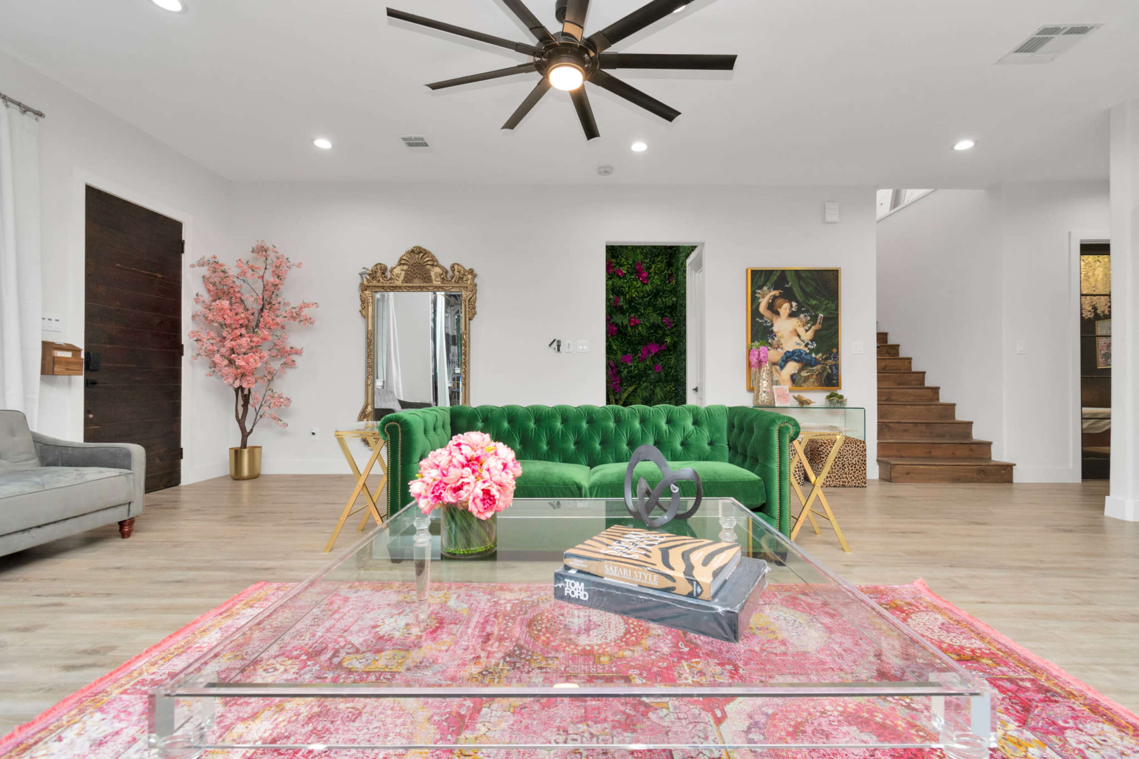 A stylish living room featuring a green velvet sofa, a large mirror, a coffee table with books, a colorful rug, and a staircase leading to an upper level.