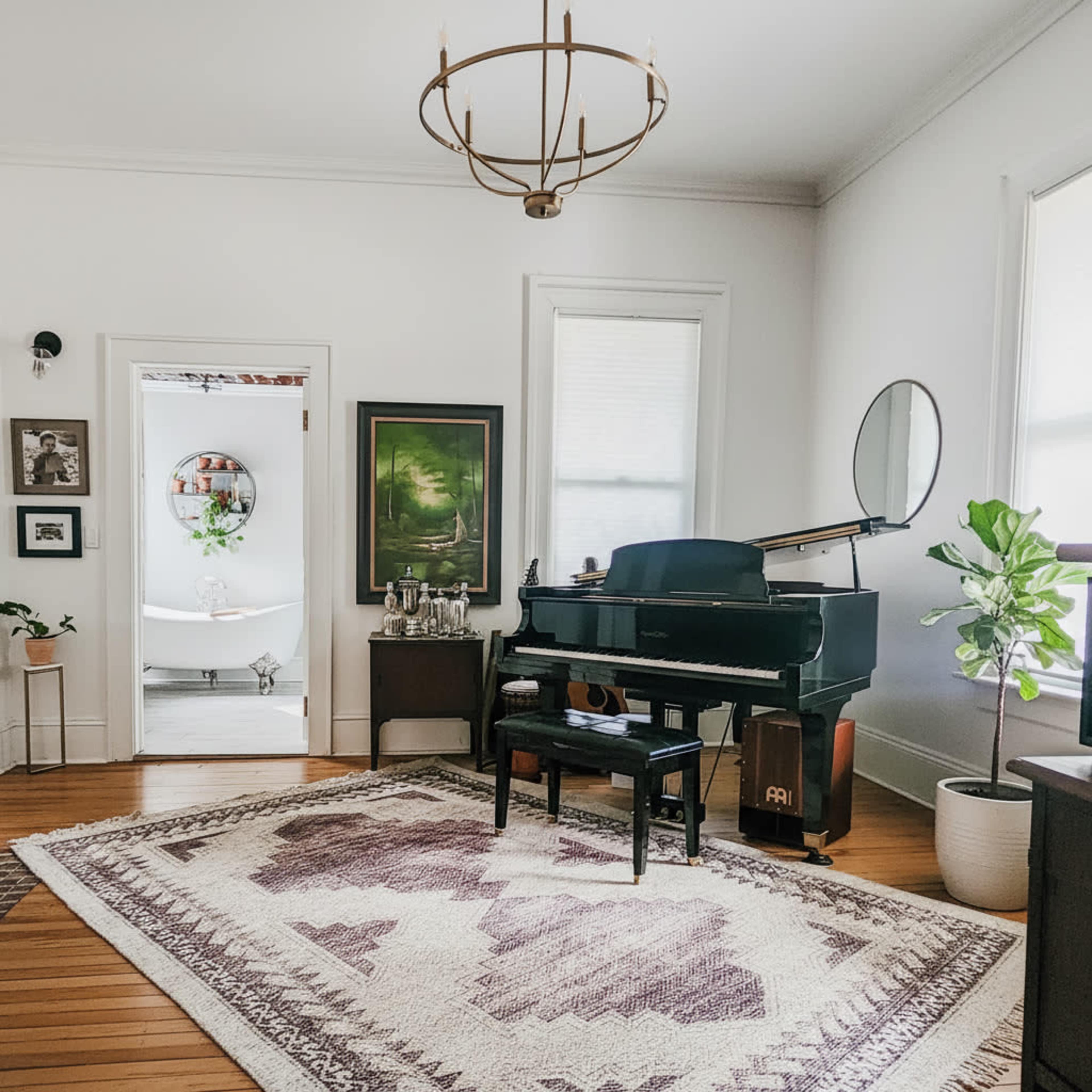 A piano and a stool are positioned in a bright room adorned with a large rug, framed artwork, and a plant.