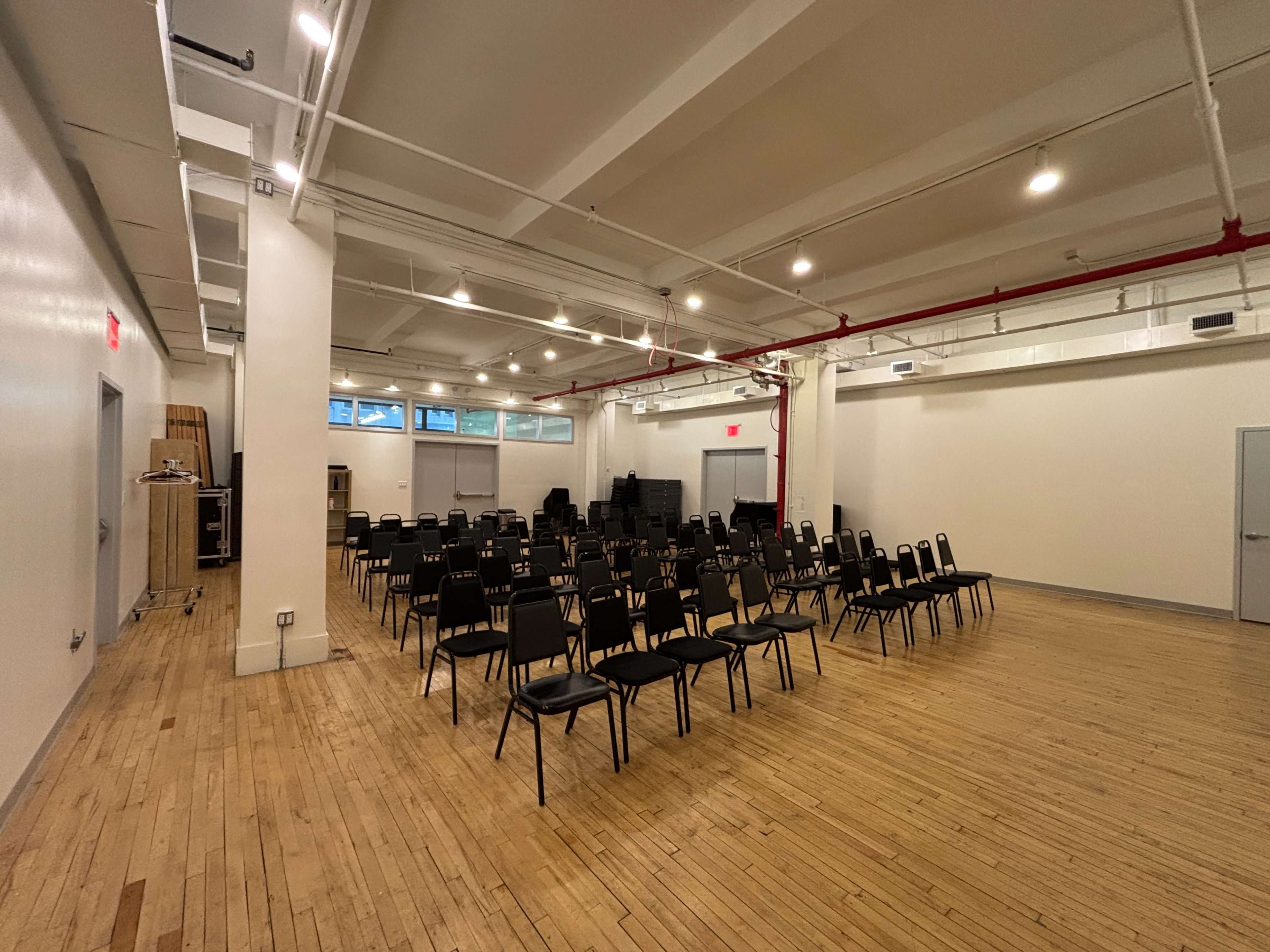 The image shows a large, empty room with neatly arranged black chairs facing a front wall, featuring wooden flooring and industrial-style lighting.