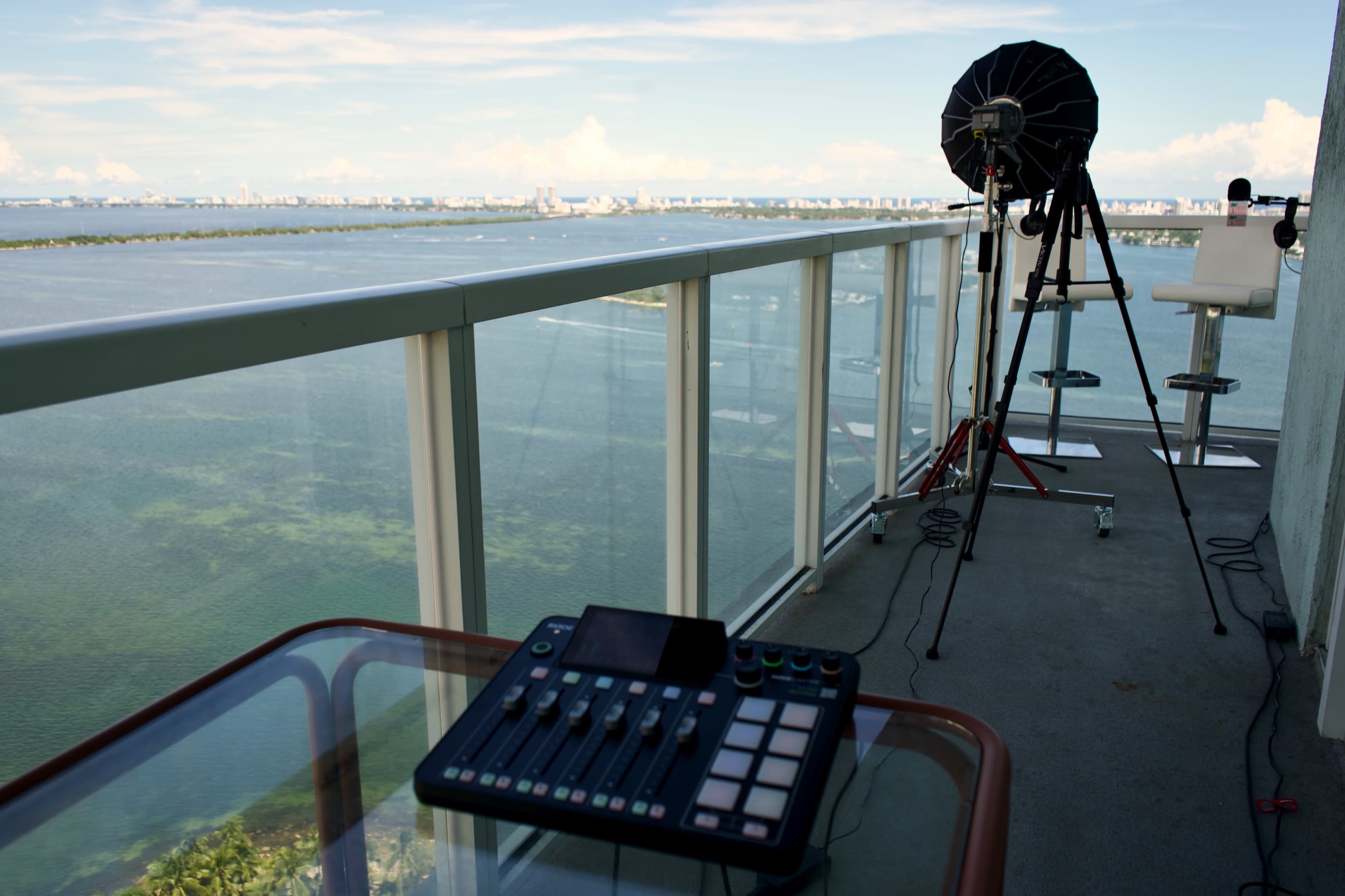 A sound mixing console is positioned on a balcony overlooking a body of water and a distant skyline.