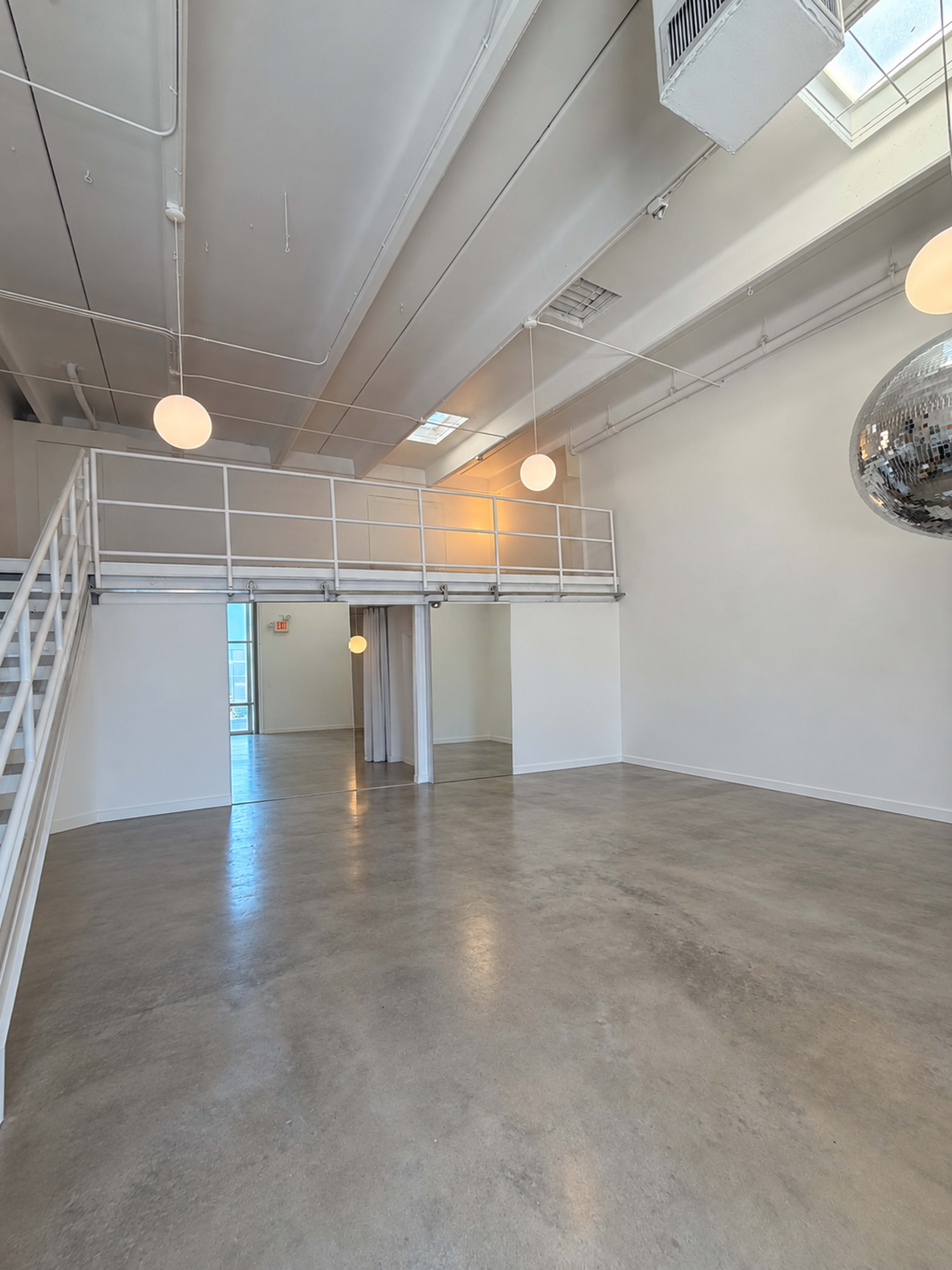 The image depicts a spacious, modern interior with a mezzanine level, large windows, and a disco ball hanging from the ceiling.