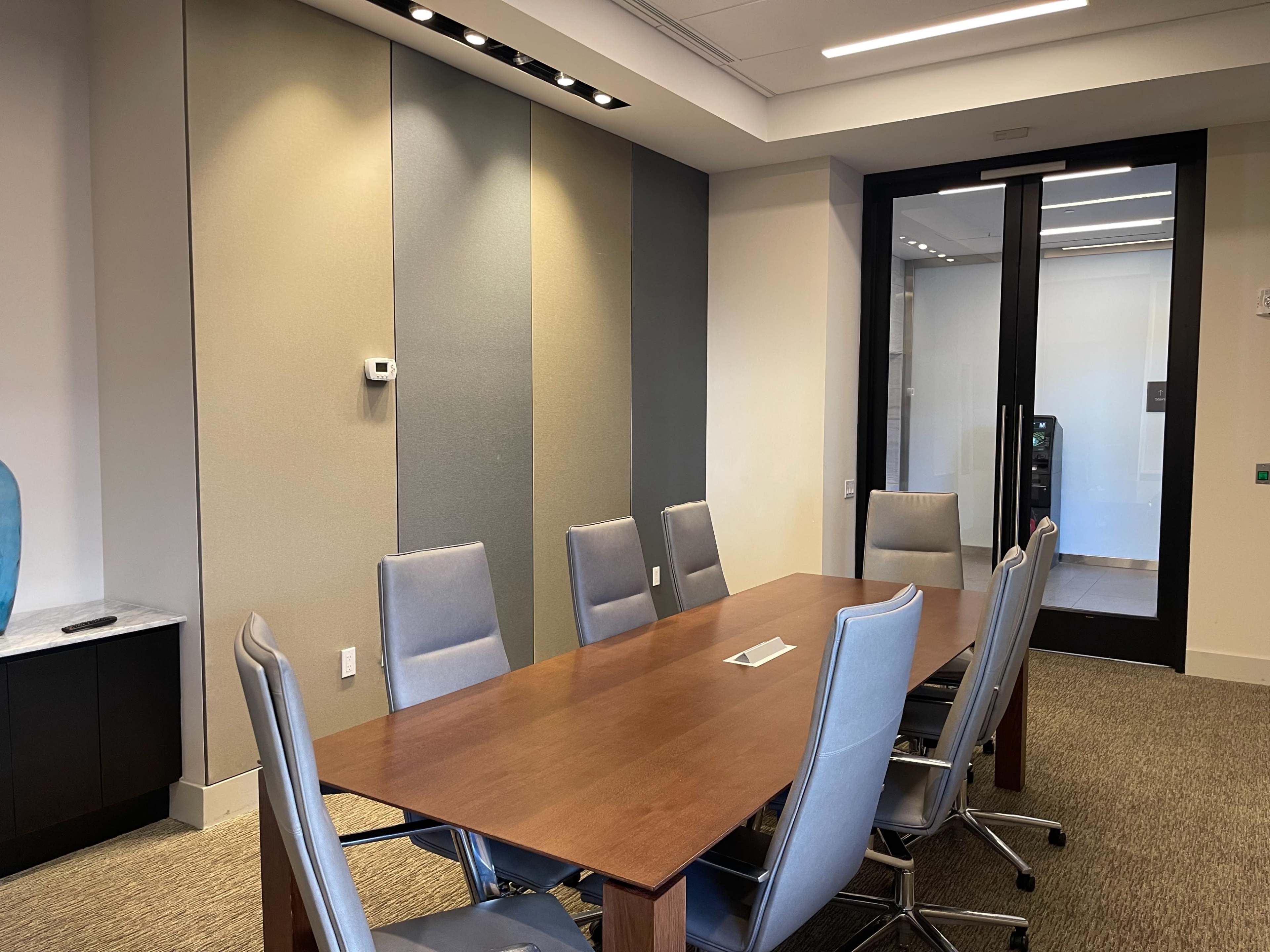 A conference room features a long wooden table surrounded by gray chairs, with a glass door leading to another area.