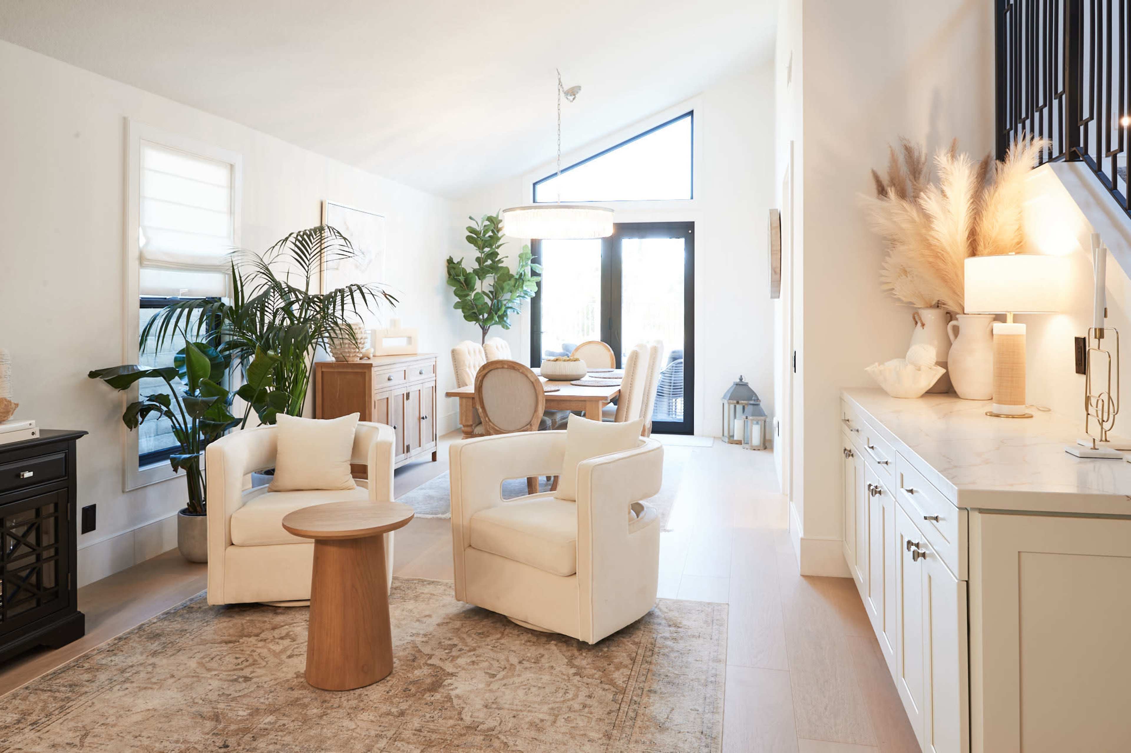 A modern living room with two white armchairs, a wooden side table, a dining area in the background, and large windows that provide natural light.