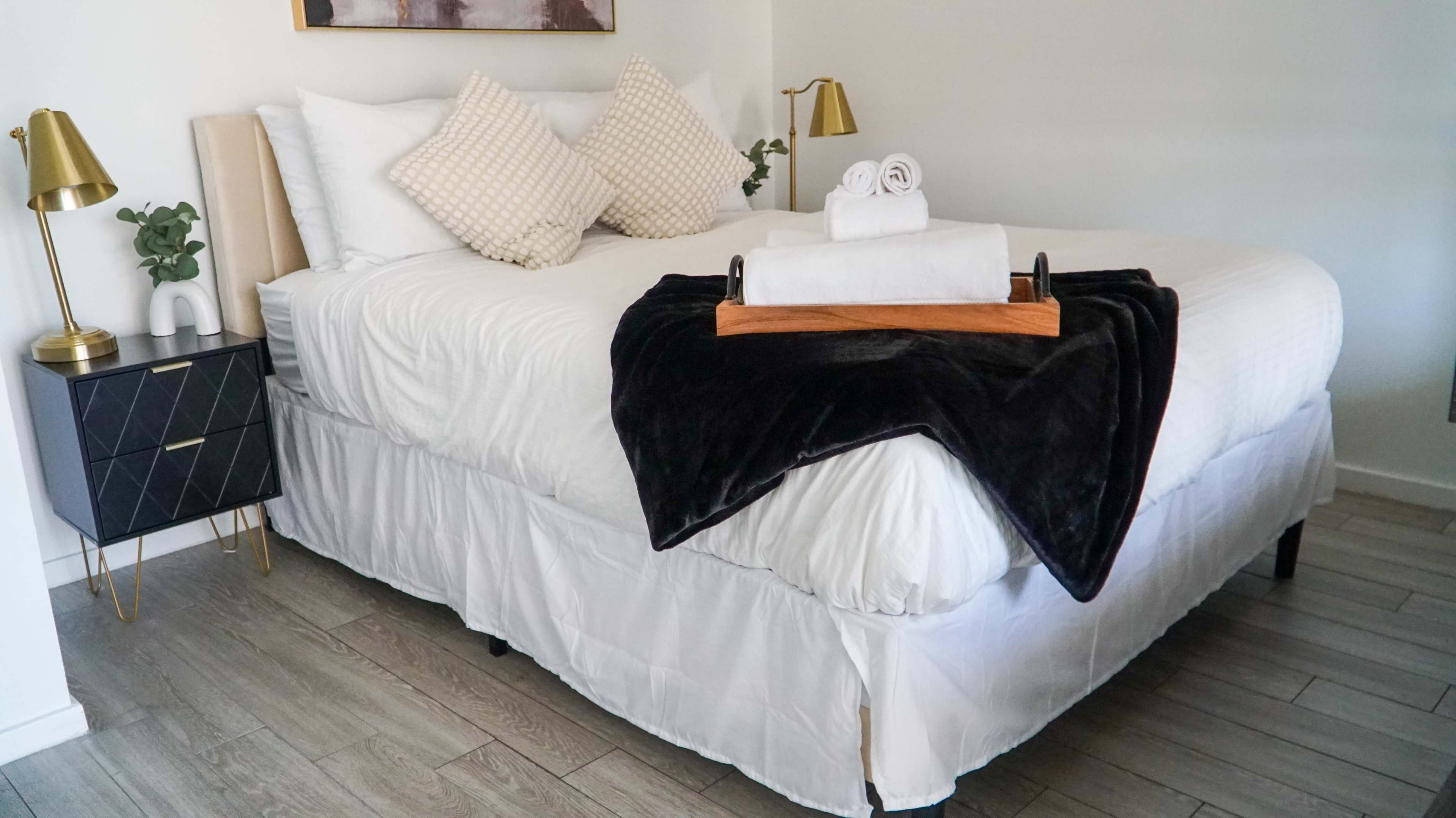 The image shows a neatly made bed with decorative pillows, a tray with rolled towels, and bedside lamps in a well-lit room.