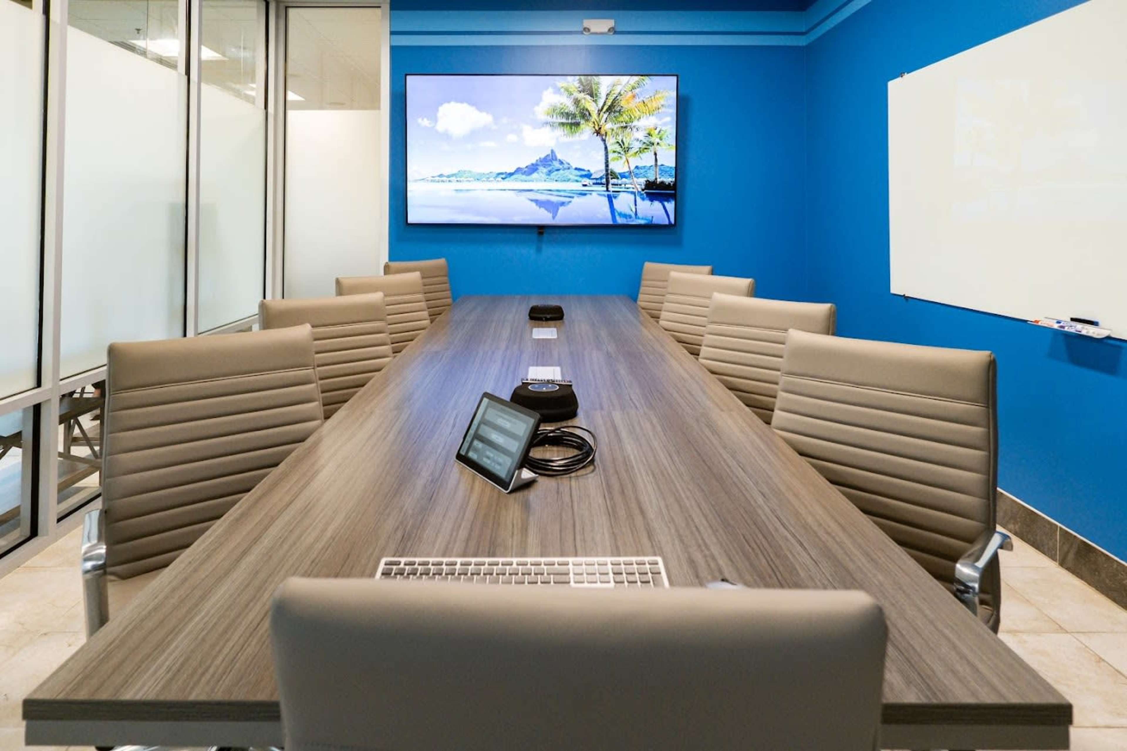 The image shows a modern conference room with a long wooden table surrounded by gray ergonomic chairs and a large television mounted on a blue wall.