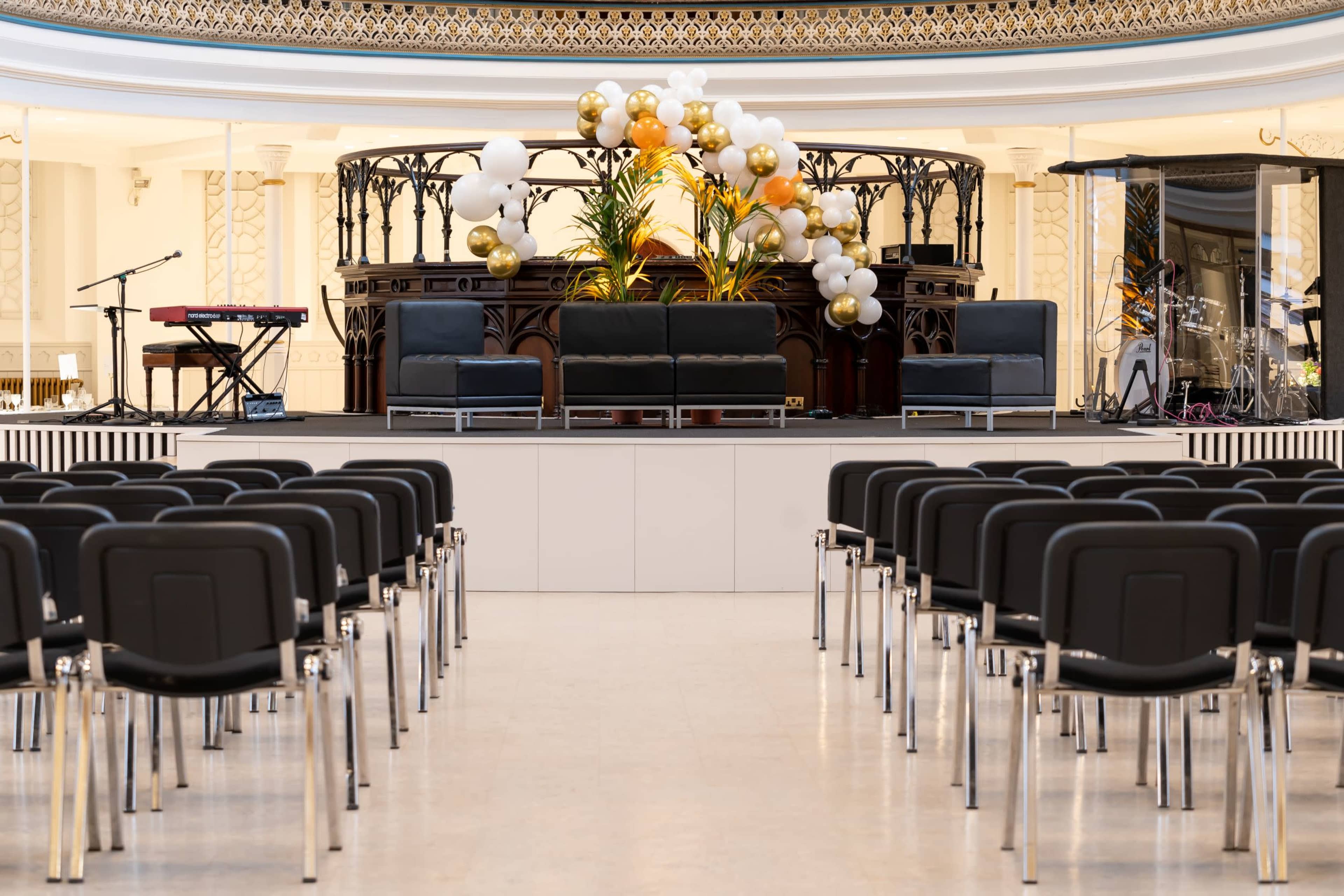 A stage with decorative balloons and plants is set up in a spacious hall, facing rows of black chairs.