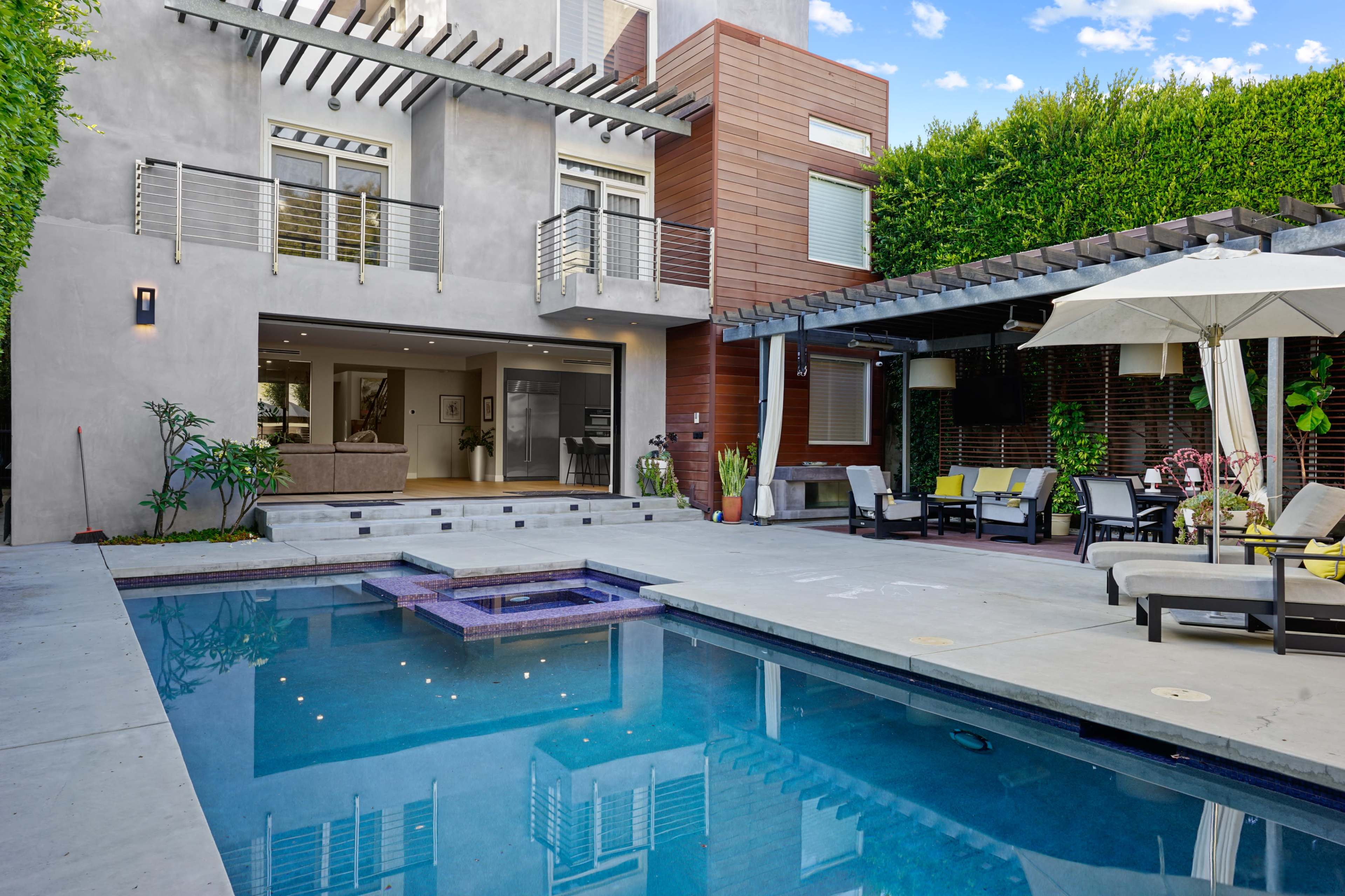 The image shows a modern backyard featuring a swimming pool with a spa, surrounded by lounge chairs and a covered seating area, alongside a contemporary house.