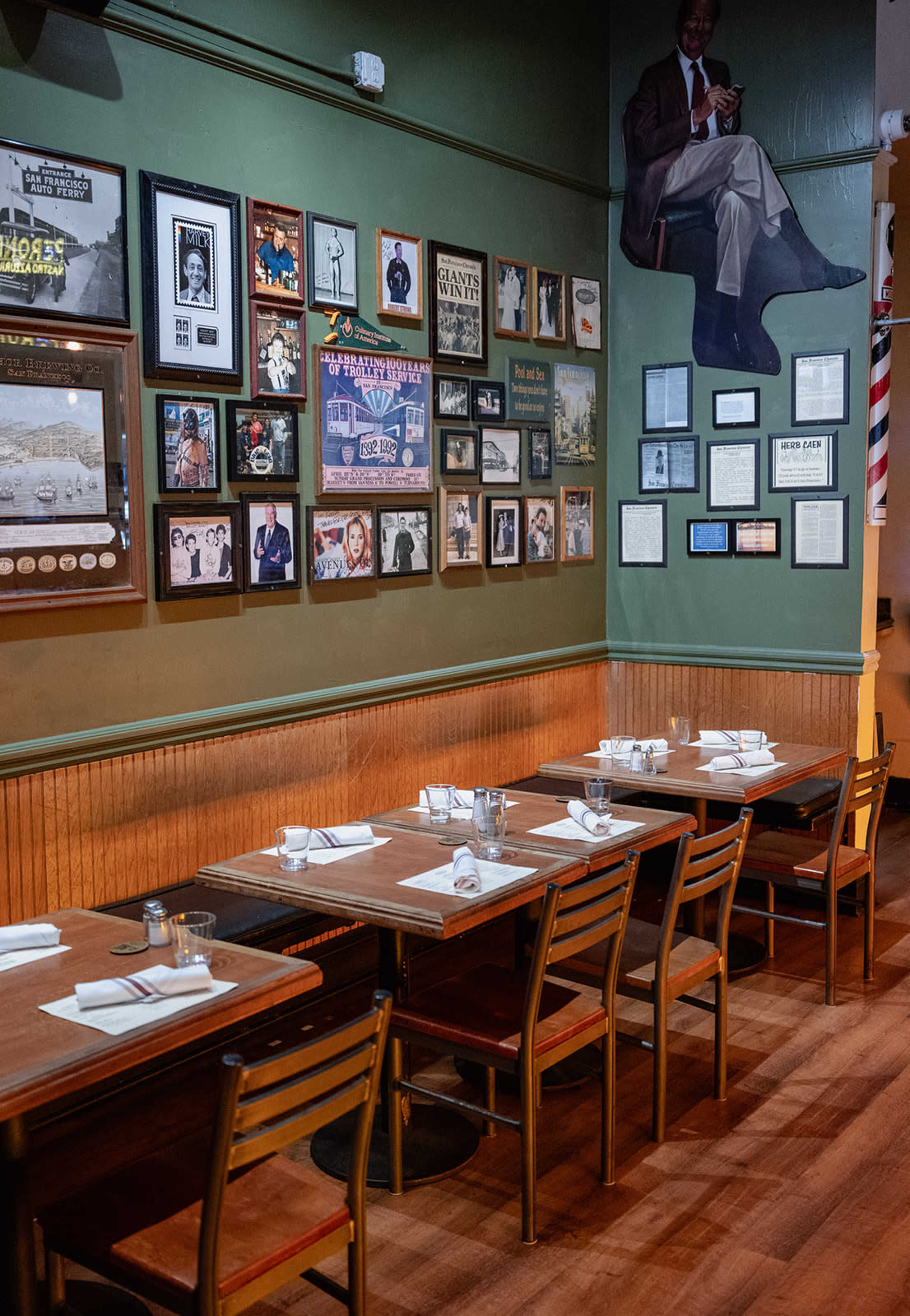 The image shows a dining area with several wooden tables set with napkins and utensils, adjacent to a wall adorned with framed photographs and documents.
