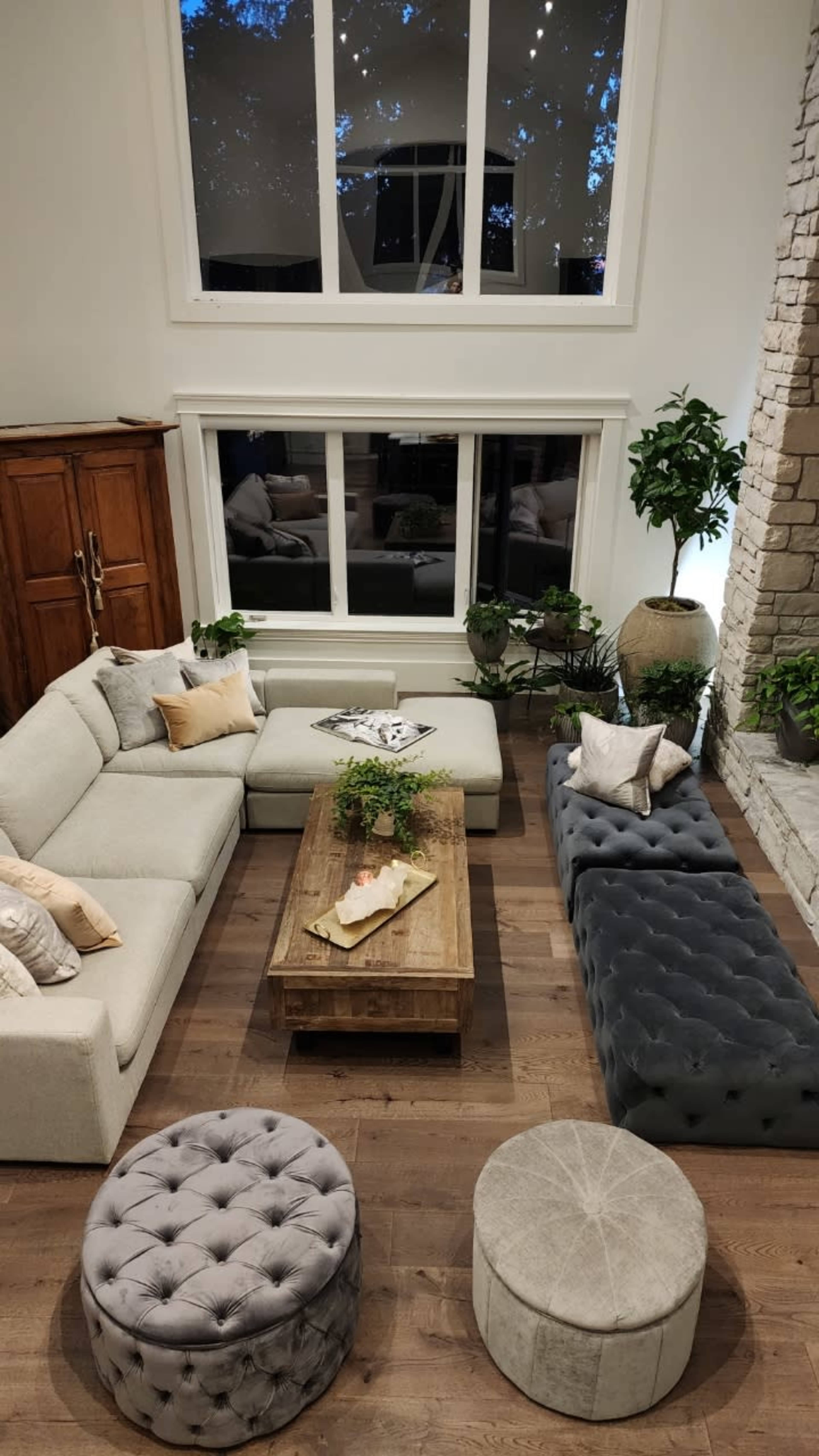 A spacious living room features a light-colored sectional sofa, a wooden coffee table, and tufted ottomans, surrounded by large windows and indoor plants.
