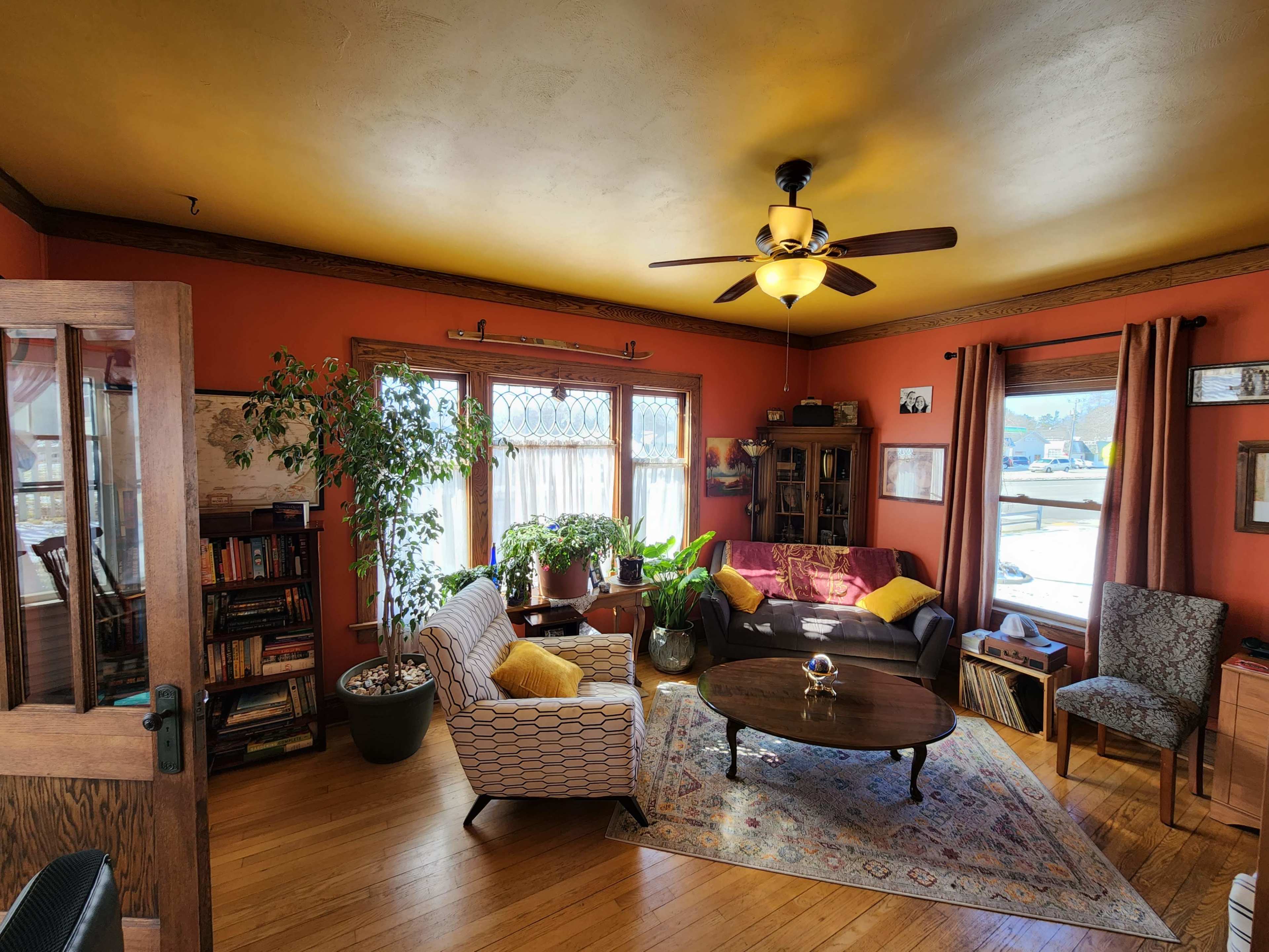 A cozy living room with an orange-walled decor, a ceiling fan, a mix of seating options, and large windows allowing natural light to enter.