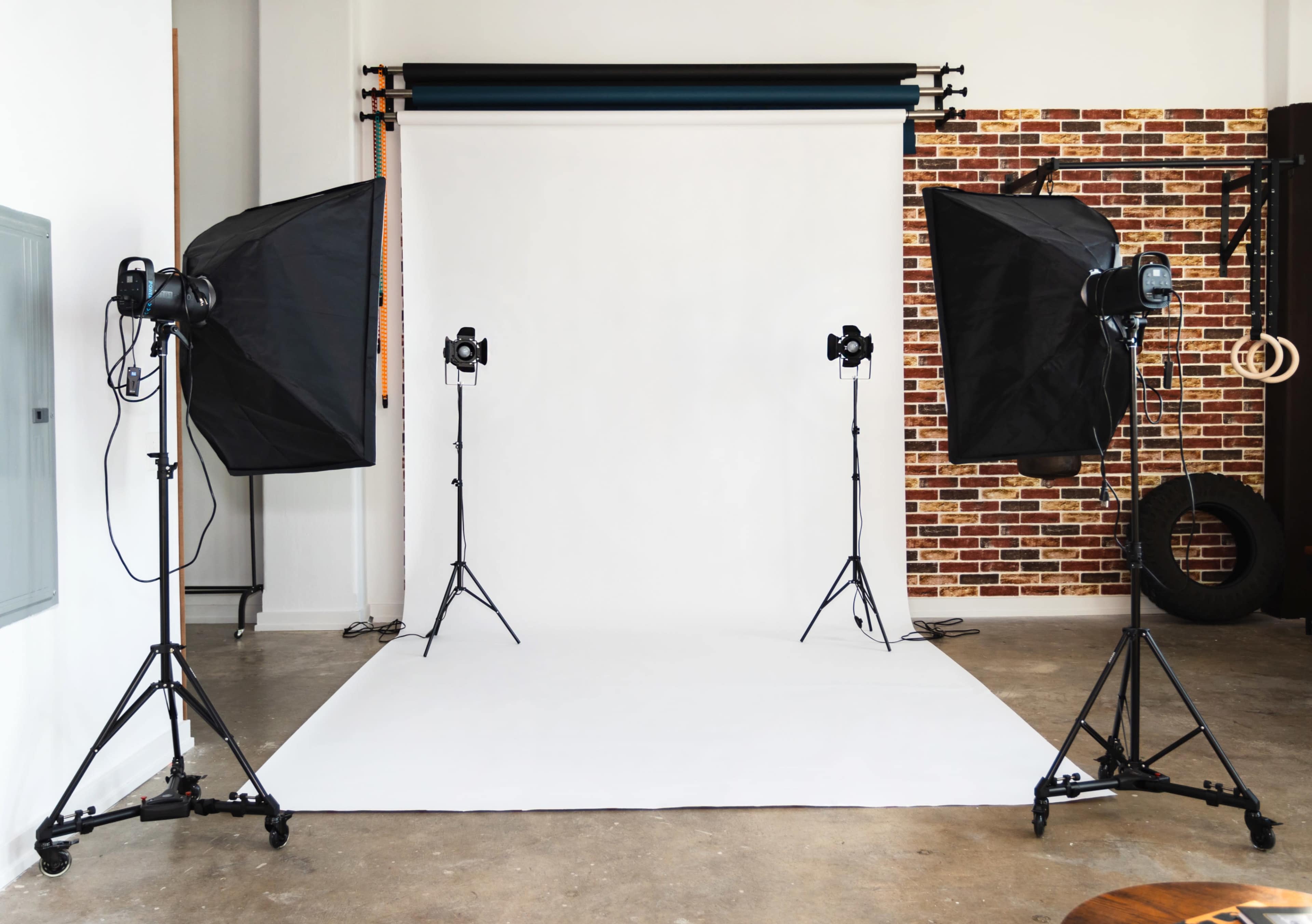 The image shows a photography studio with a white backdrop, two softbox lights on stands, and a textured brick wall in the background.
