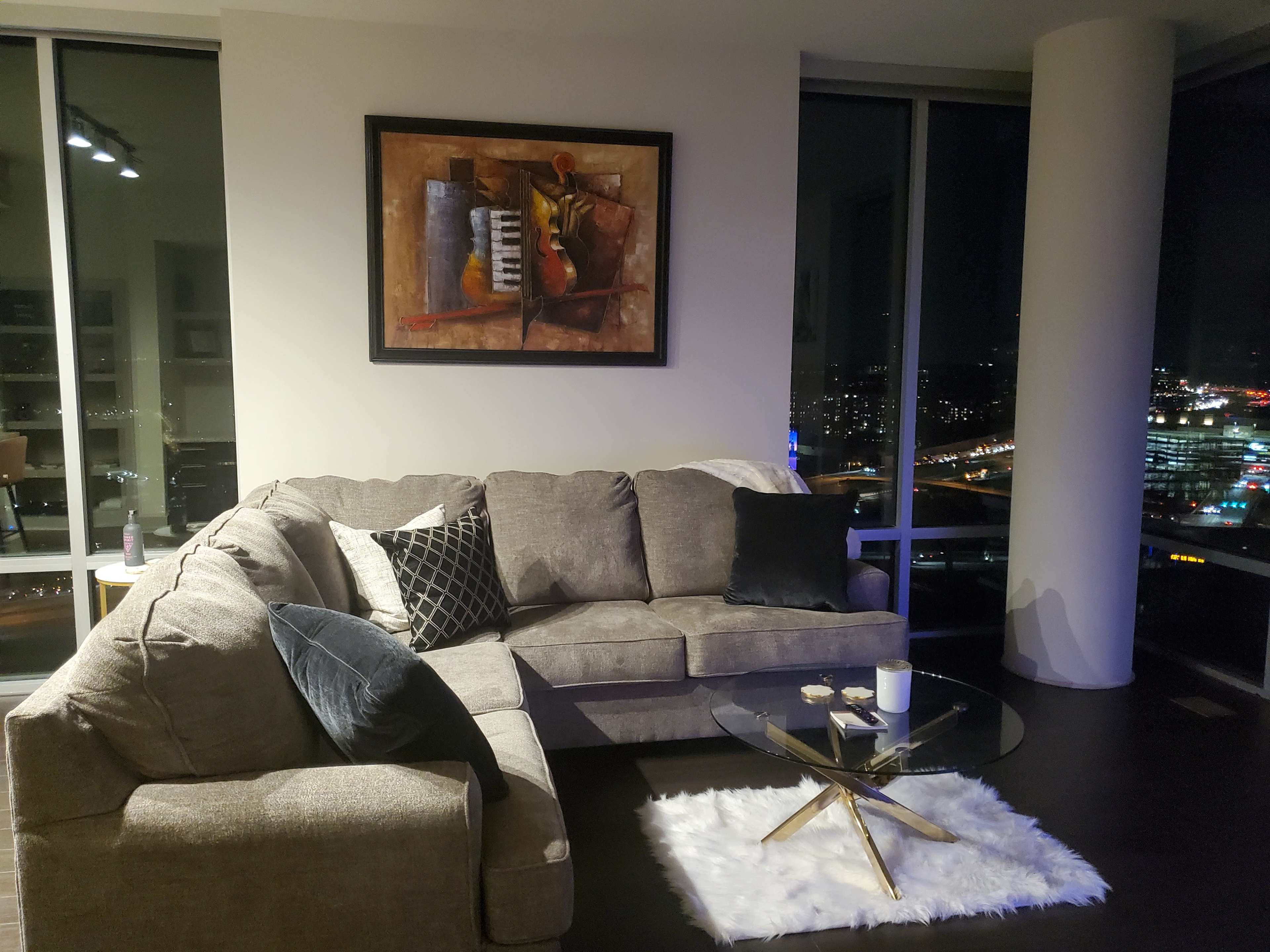 A modern living room features a grey sectional sofa, a glass coffee table on a white rug, and a framed painting on the wall, with city skyline views visible through large windows.