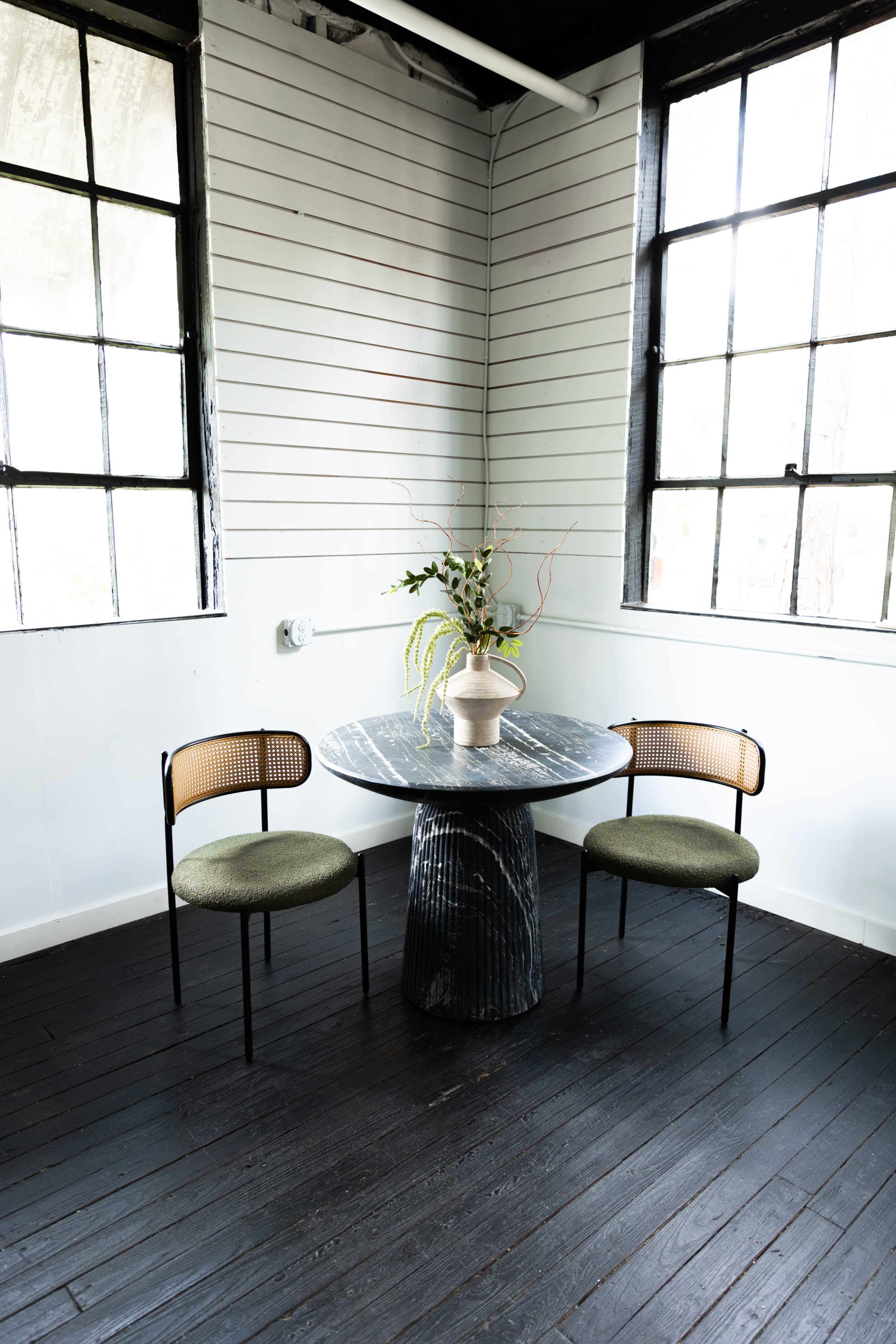 A round table with a textured base and a vase of greenery is positioned between two chairs in a well-lit corner of a room with white walls and large windows.