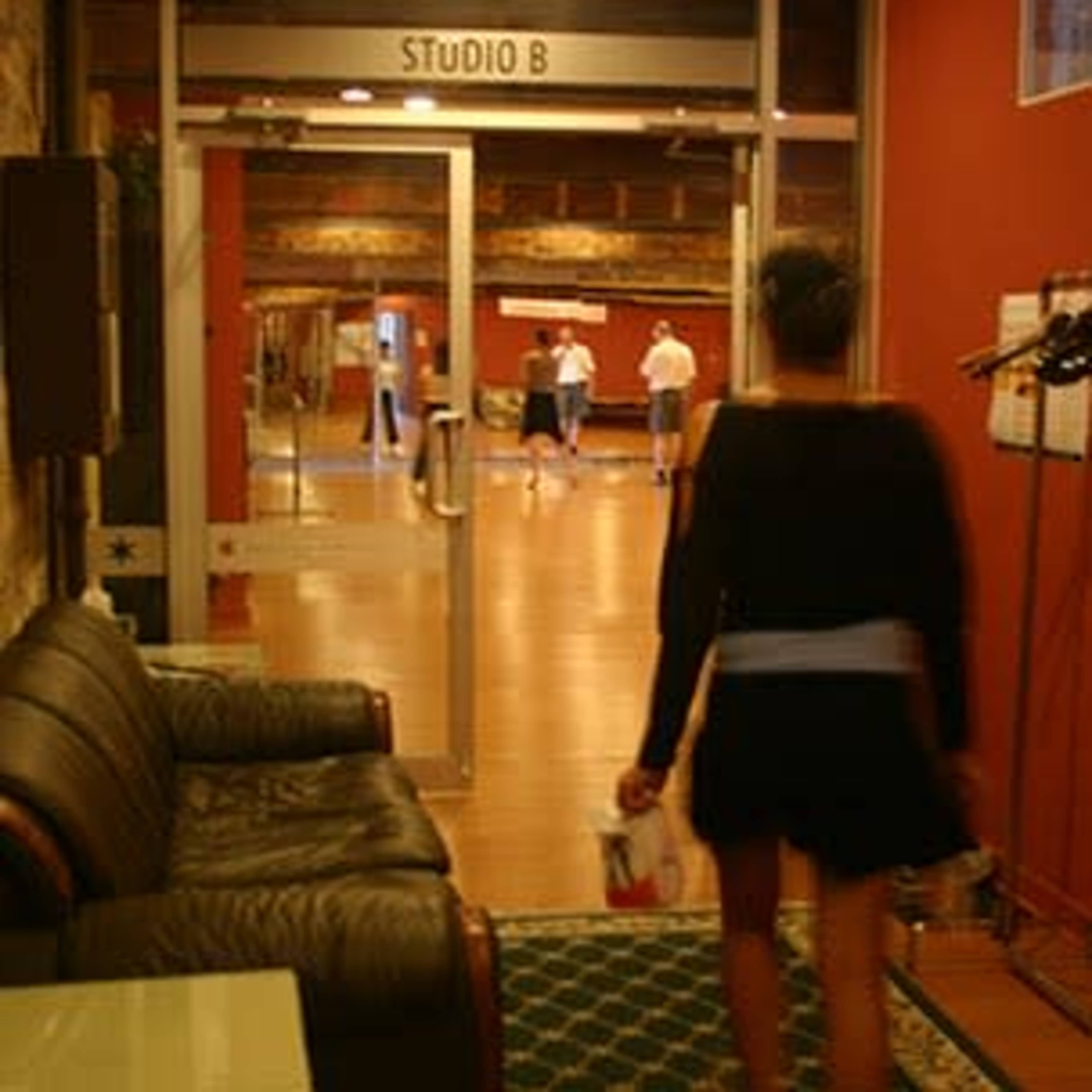 A person walks toward a dance studio entrance with a wooden floor, where others are practicing in the background.