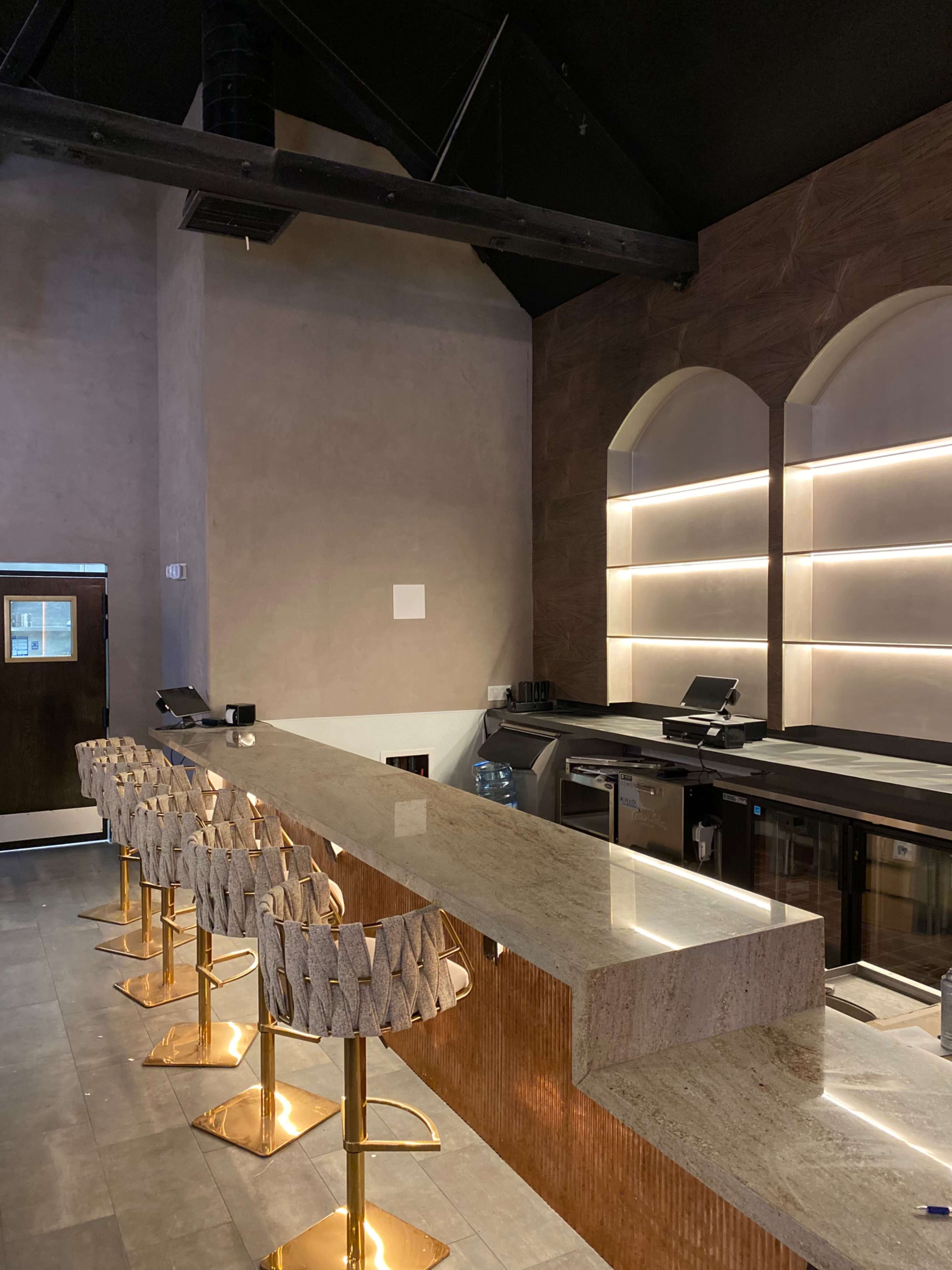 A modern bar area with a sleek marble countertop and several golden bar stools arranged in front of it, illuminated by subtle lighting.