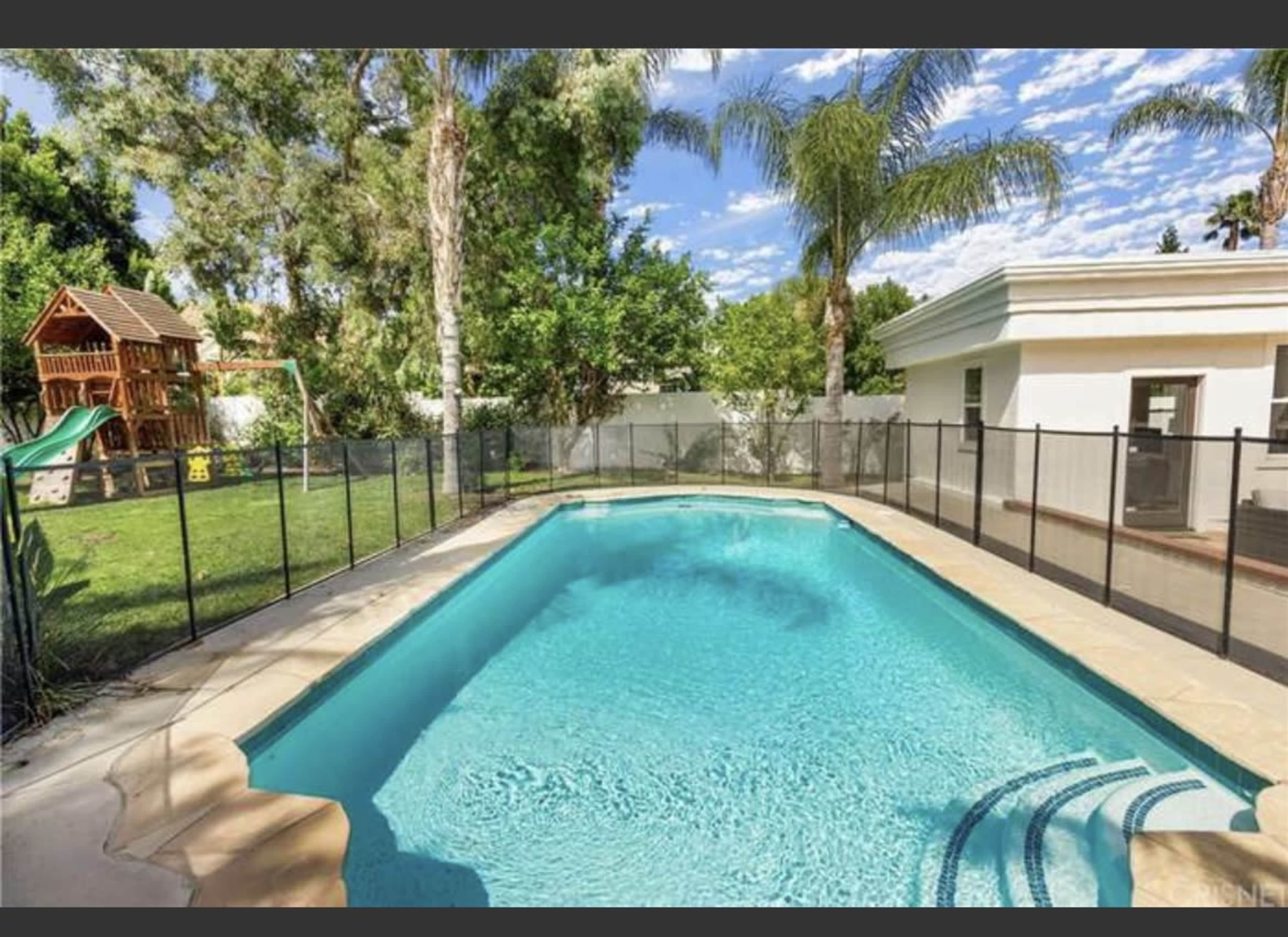A fenced swimming pool with clear water, surrounded by grass and a playground set in a residential backyard.