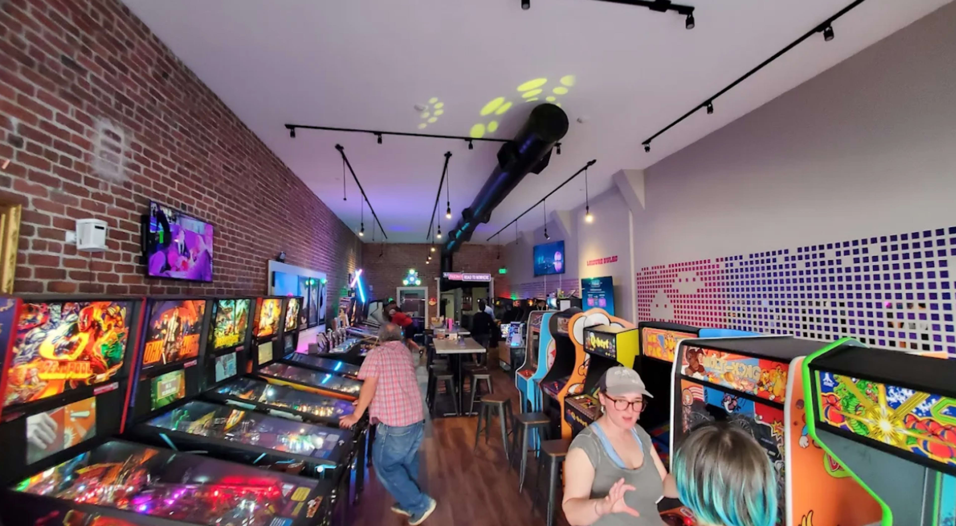 The image shows an arcade filled with various pinball machines and retro gaming consoles, with people interacting and enjoying the games in a well-lit, modern space.