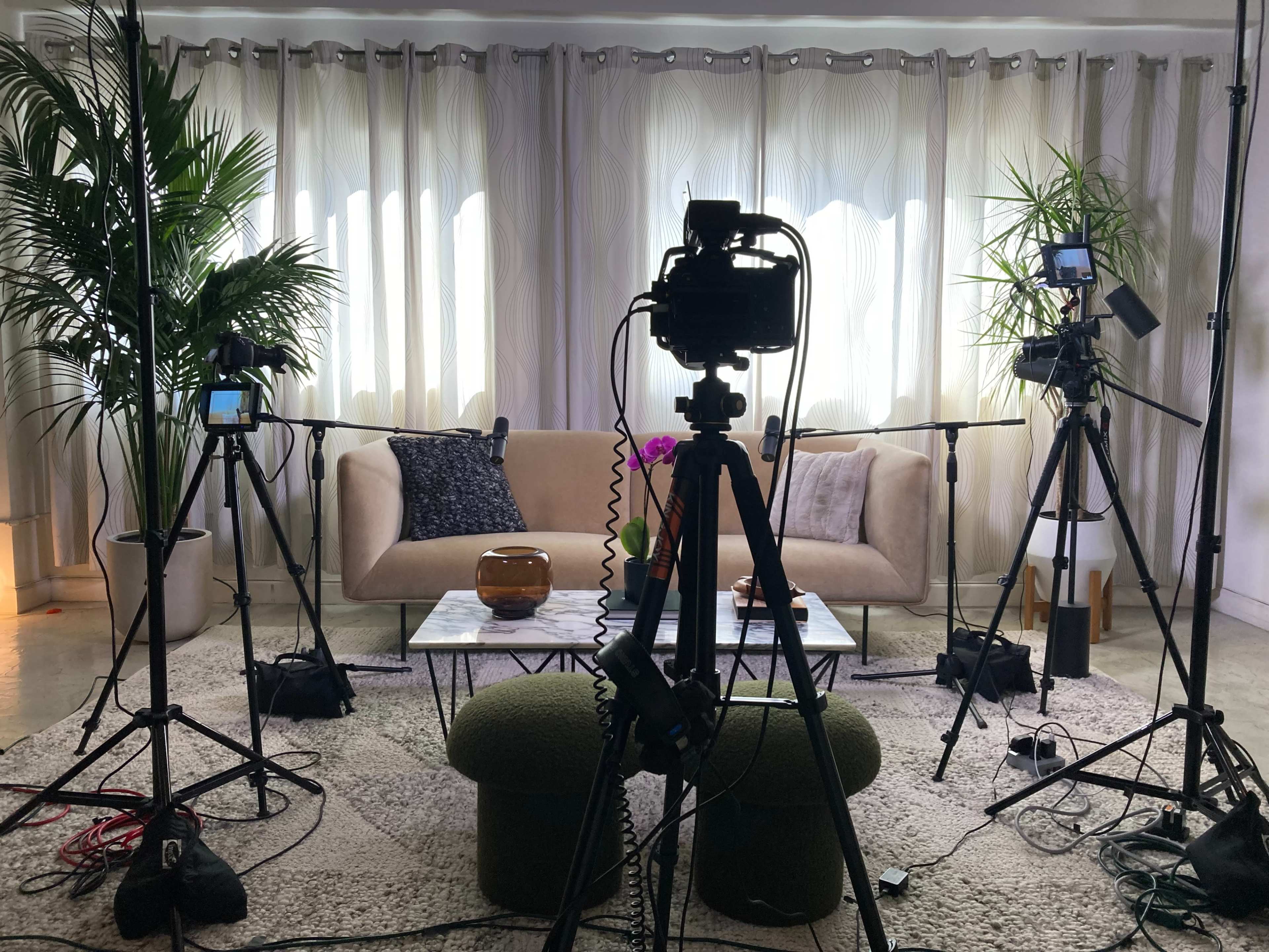 The image shows a studio setup with multiple cameras, tripods, and a sofa in front of a curtain backdrop, alongside potted plants.