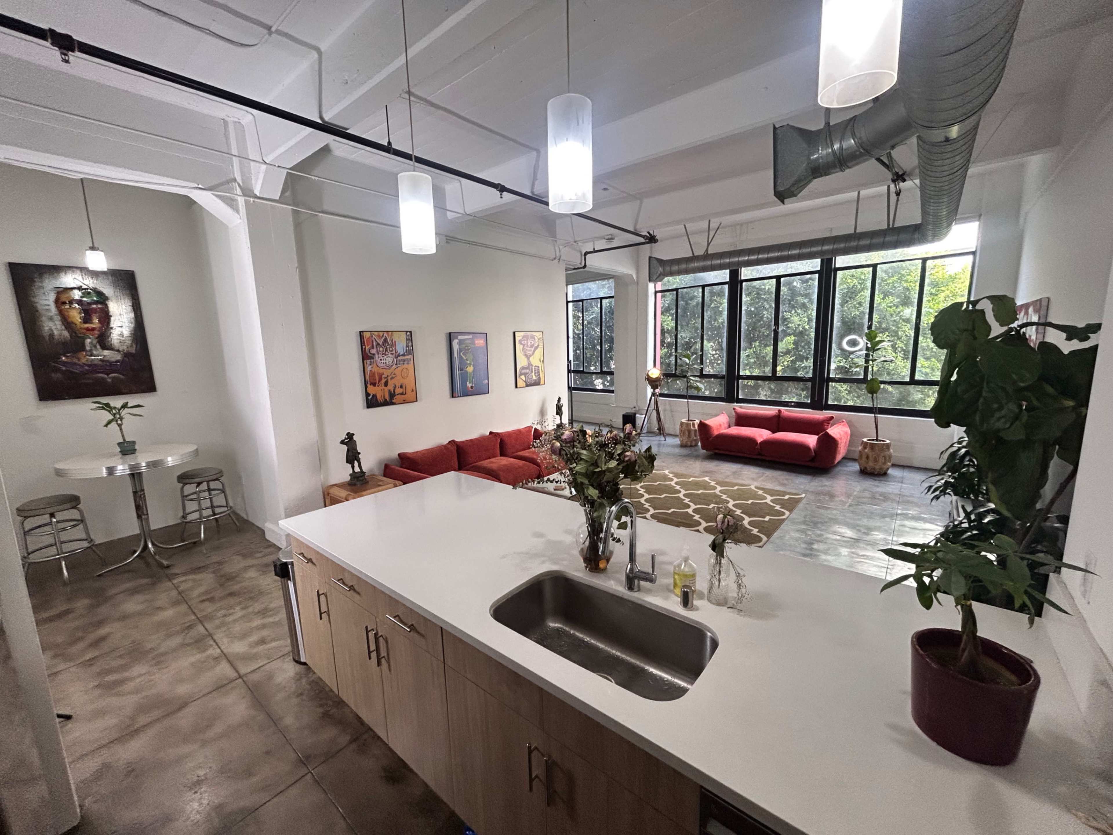 The image shows a spacious open-concept room with a kitchen area, a seating area featuring red couches, and large windows providing natural light.