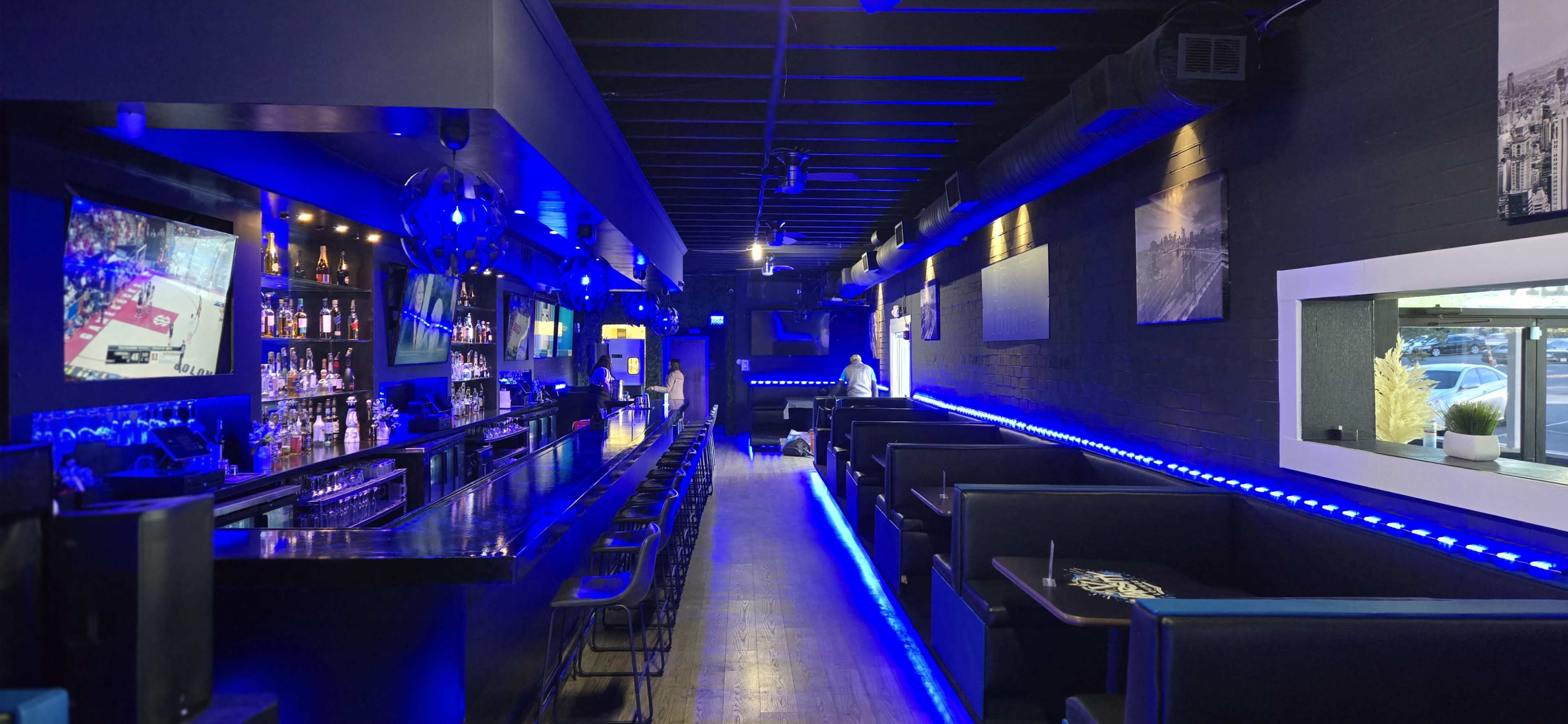 The image shows a modern bar interior featuring a long counter with bar stools, illuminated by blue LED lights, and equipped with multiple televisions displaying sports.