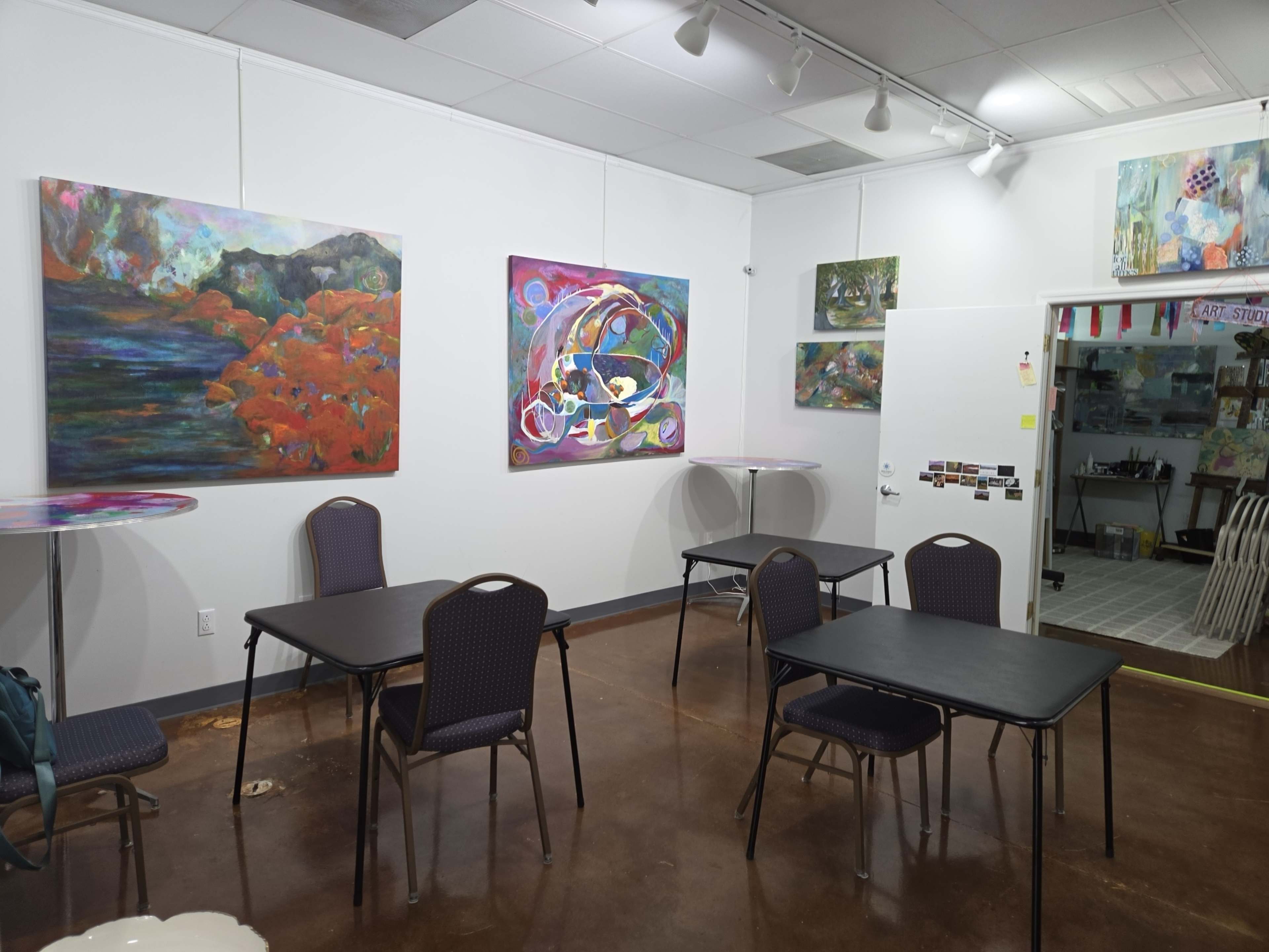 The image shows an art studio with various colorful paintings displayed on the walls and several tables and chairs arranged throughout the space.