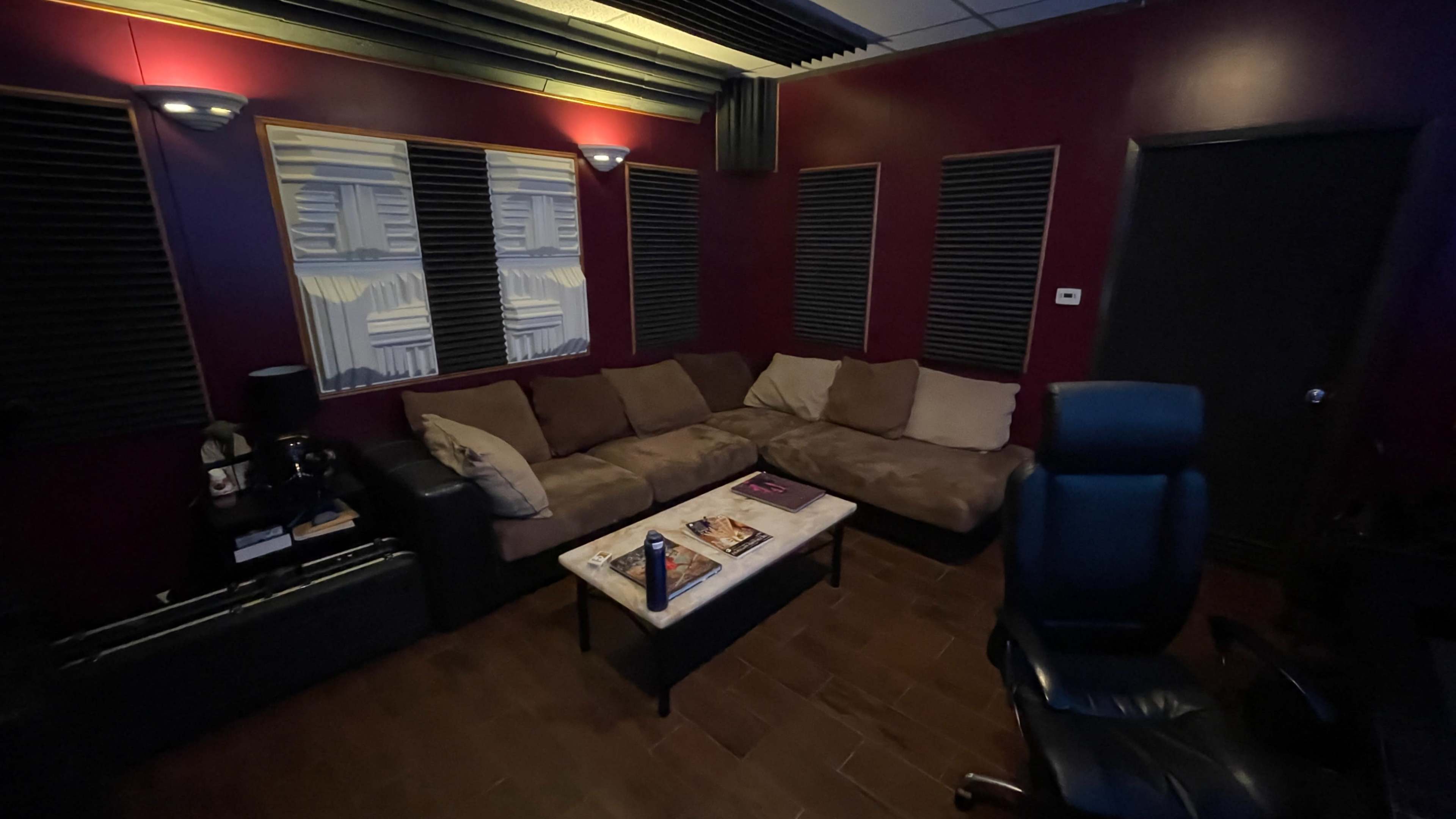 A cozy recording studio with a large brown sectional couch, a coffee table, soundproofing panels on the walls, and a black office chair.