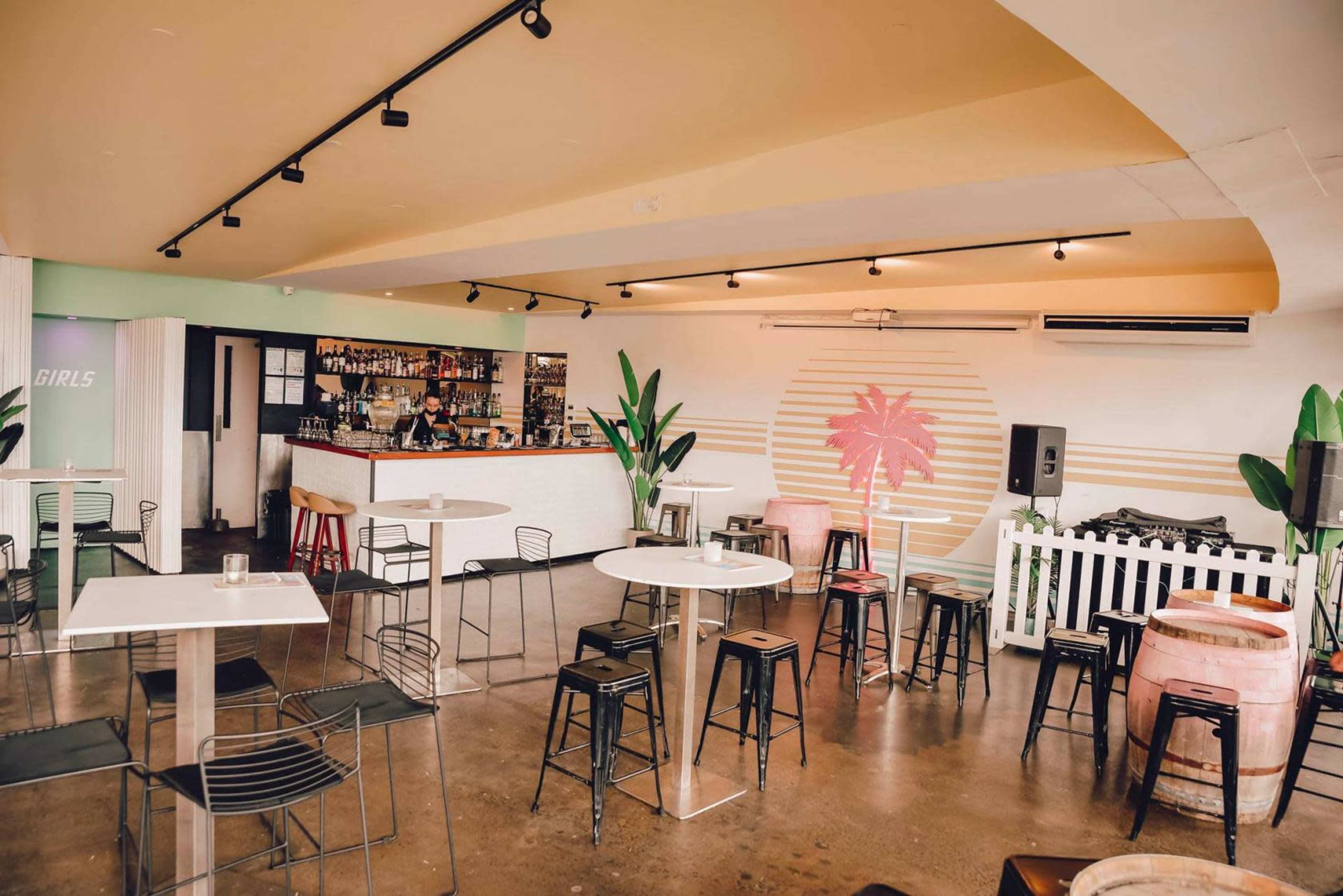 A modern bar area with a light-colored interior, featuring several tables and chairs, a bar counter stocked with drinks, and tropical-themed decor on the walls.