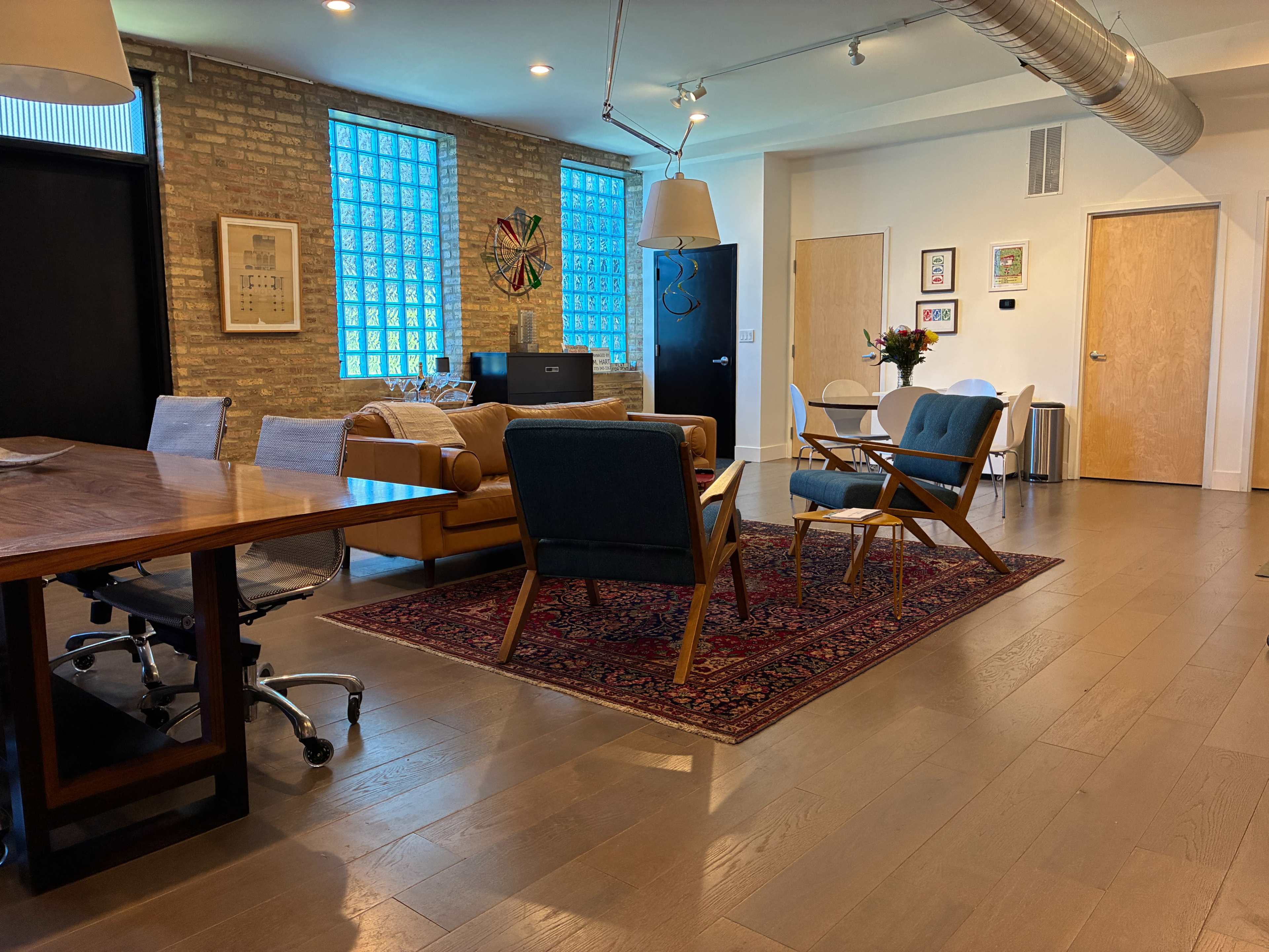 The image shows a modern interior space featuring a dining table, comfortable seating, a rug, and large windows with natural light.