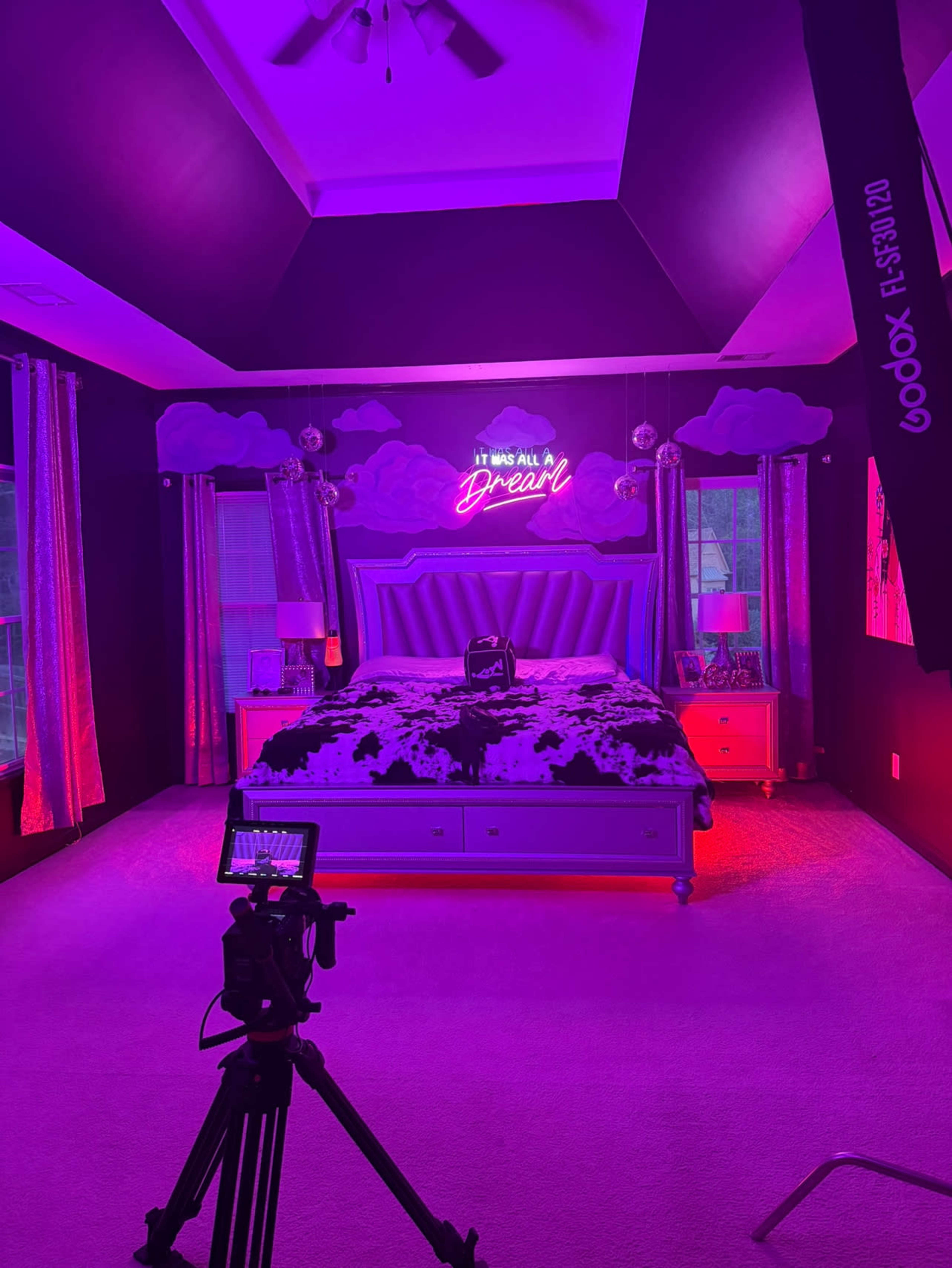 A brightly lit bedroom features a purple color scheme, a large bed with a cow-print blanket, and a neon sign that reads "It was all a dream."