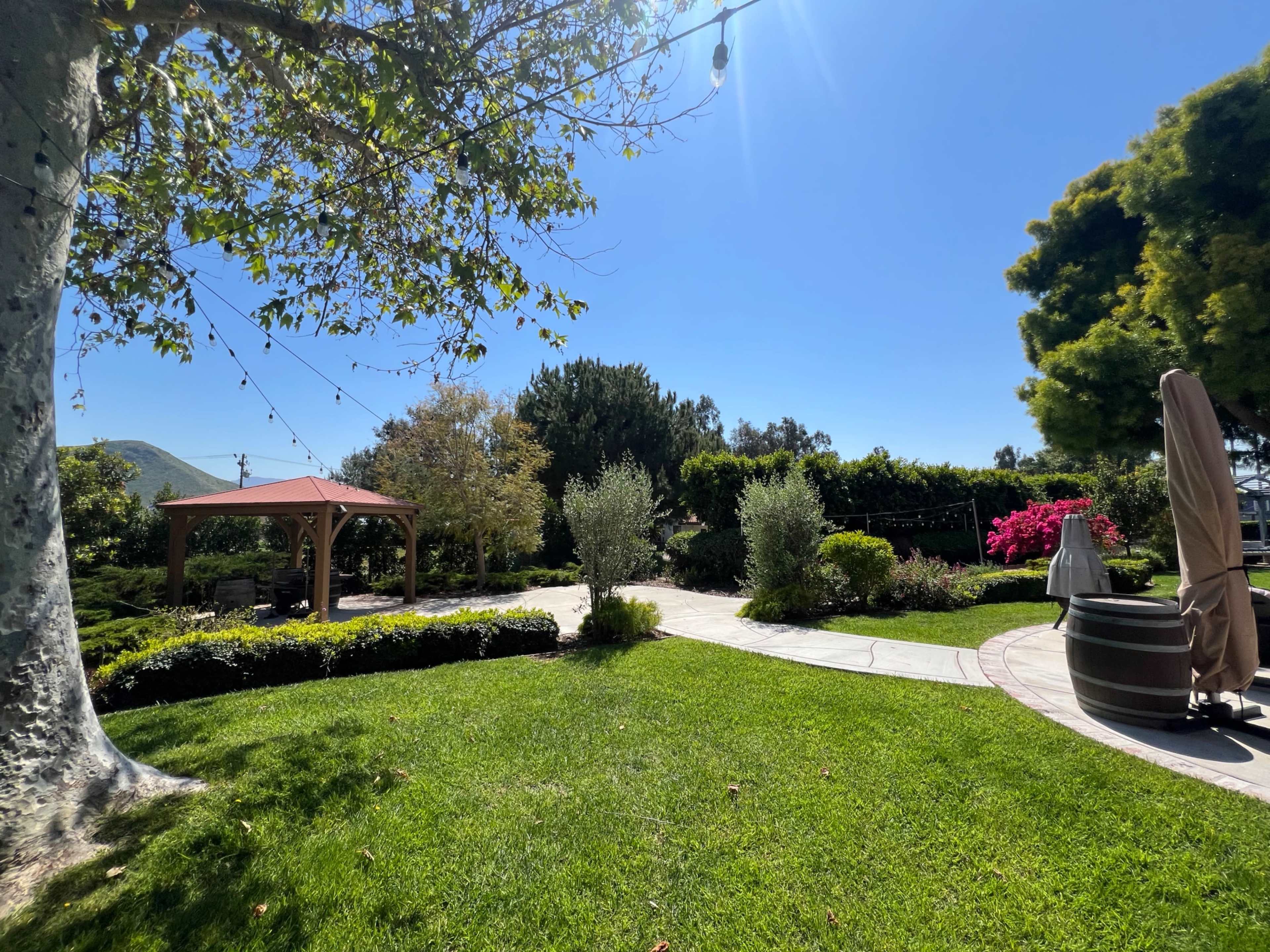 The image shows a landscaped garden with a gazebo, neatly trimmed grass, flower beds, and a stone pathway under a clear blue sky.