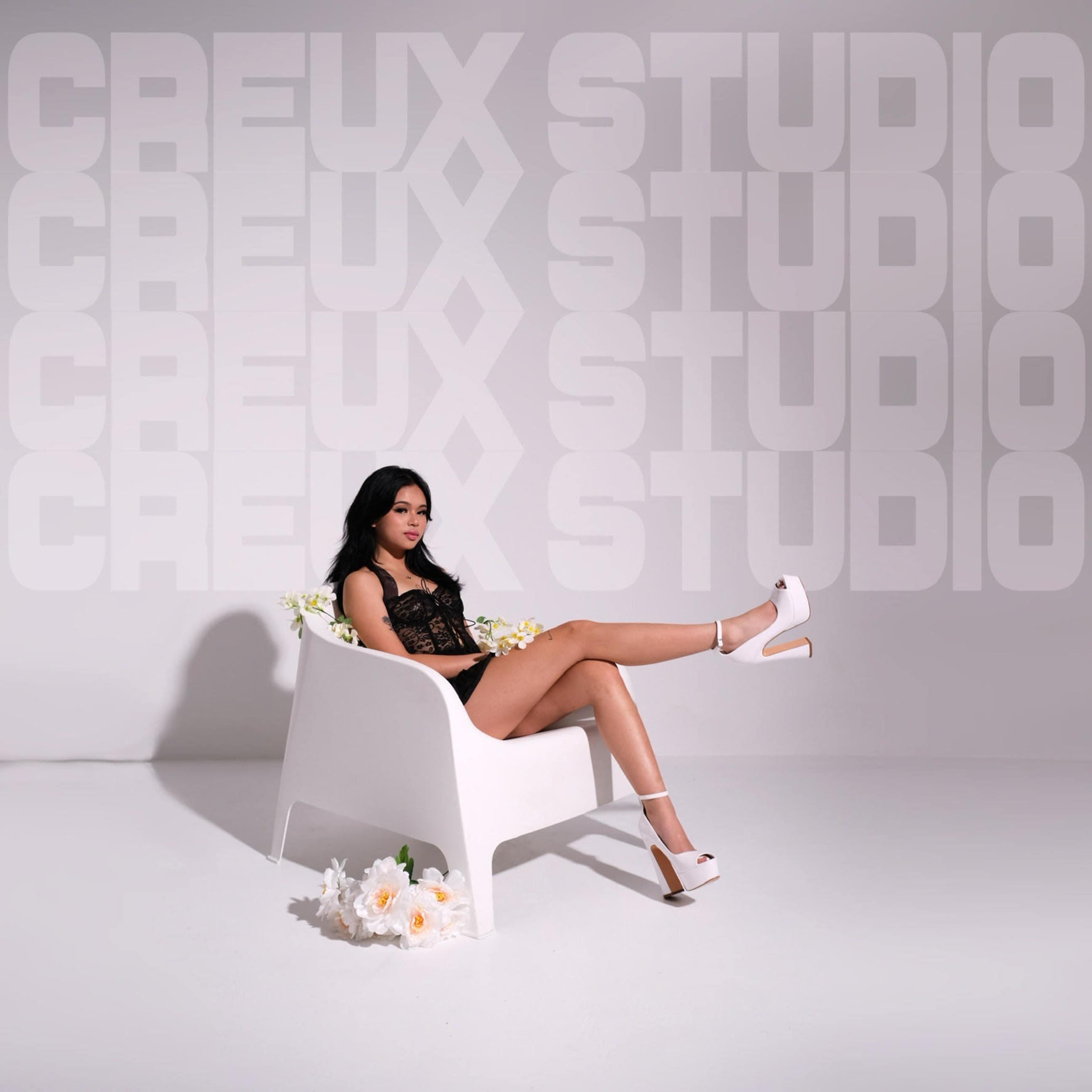 A woman sits in a white chair with her legs extended, surrounded by flowers and a backdrop featuring the text "CREUX STUDIO."