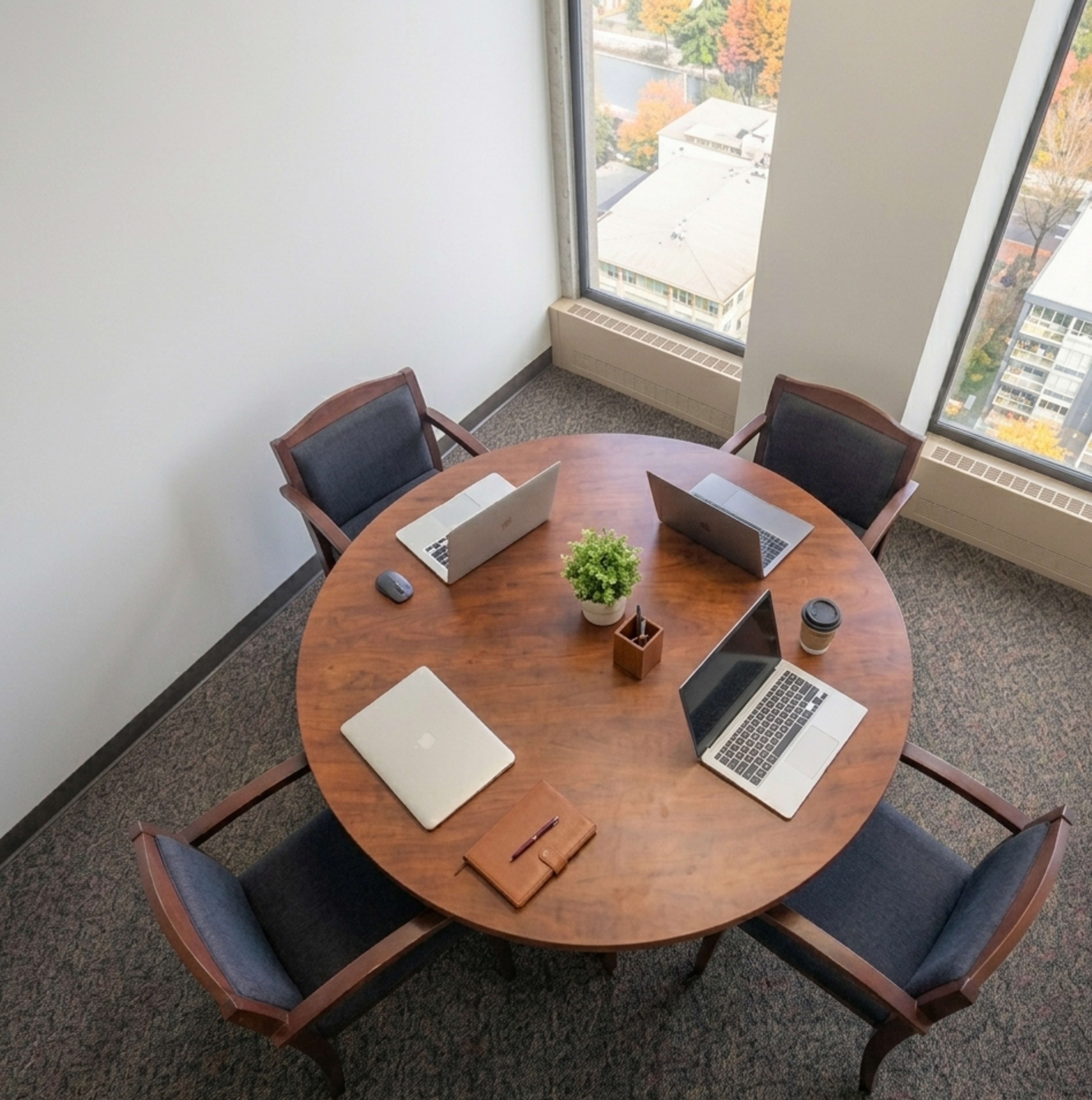 A round wooden table is set with four laptops, a small plant, a coffee cup, and two chairs in a well-lit office space with large windows.