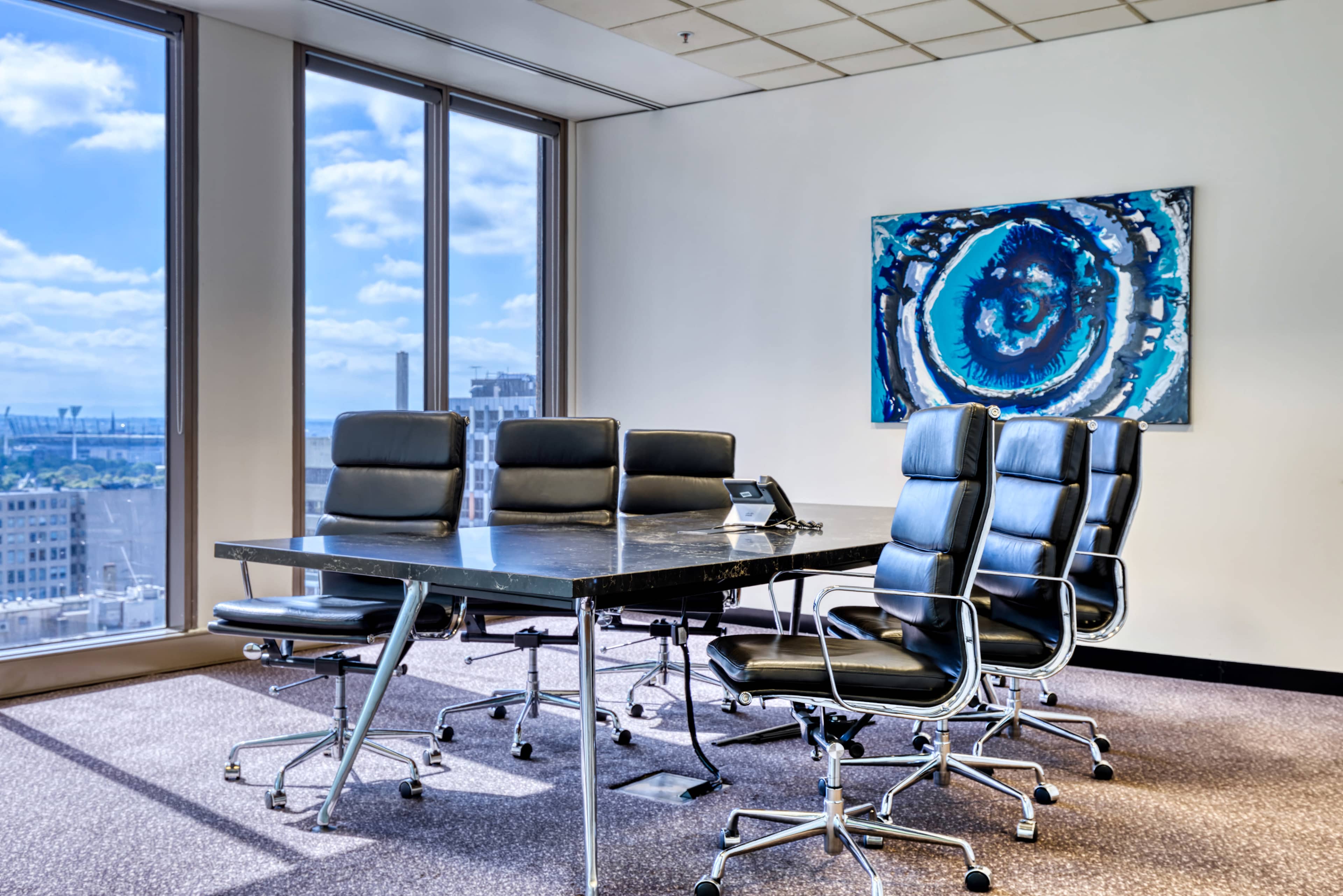 A modern conference room features a large table surrounded by black leather chairs and a colorful abstract painting on the wall.