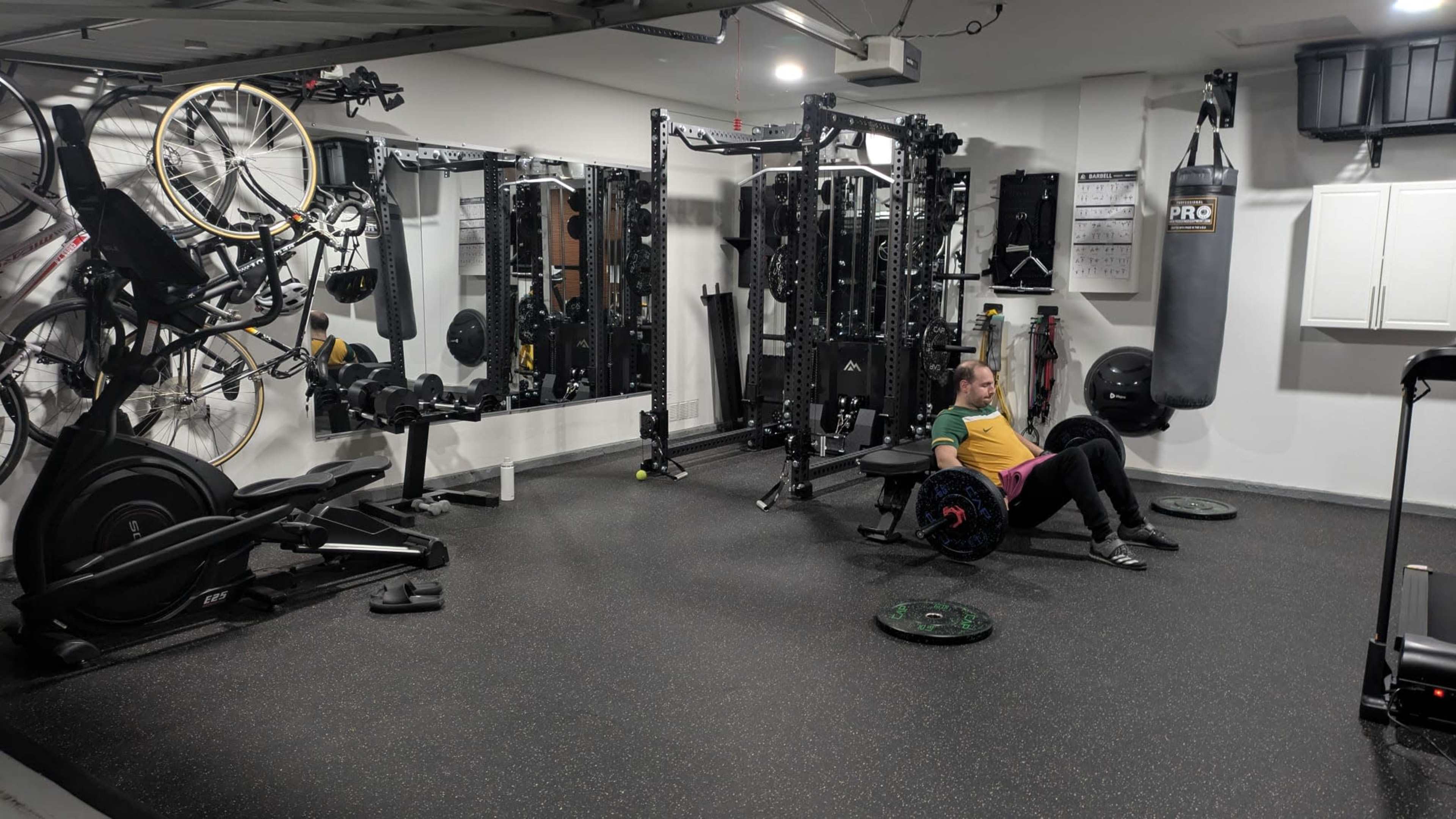 The image shows a home gym equipped with various fitness machines, weights, and a person seated on a weight bench.