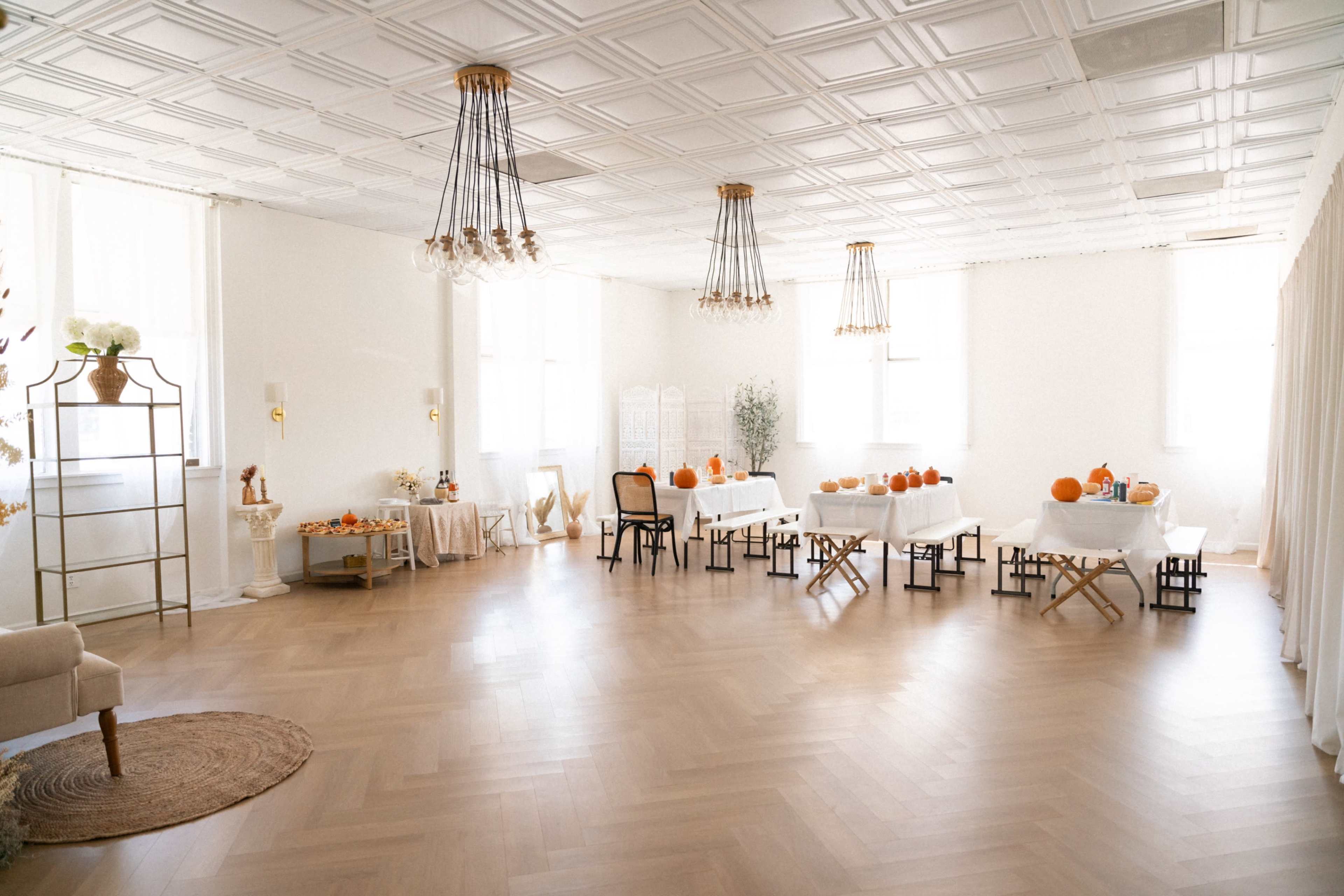 A spacious, well-lit room features several tables adorned with pumpkins and decorative items, surrounded by neutral-colored furnishings and a wooden floor.