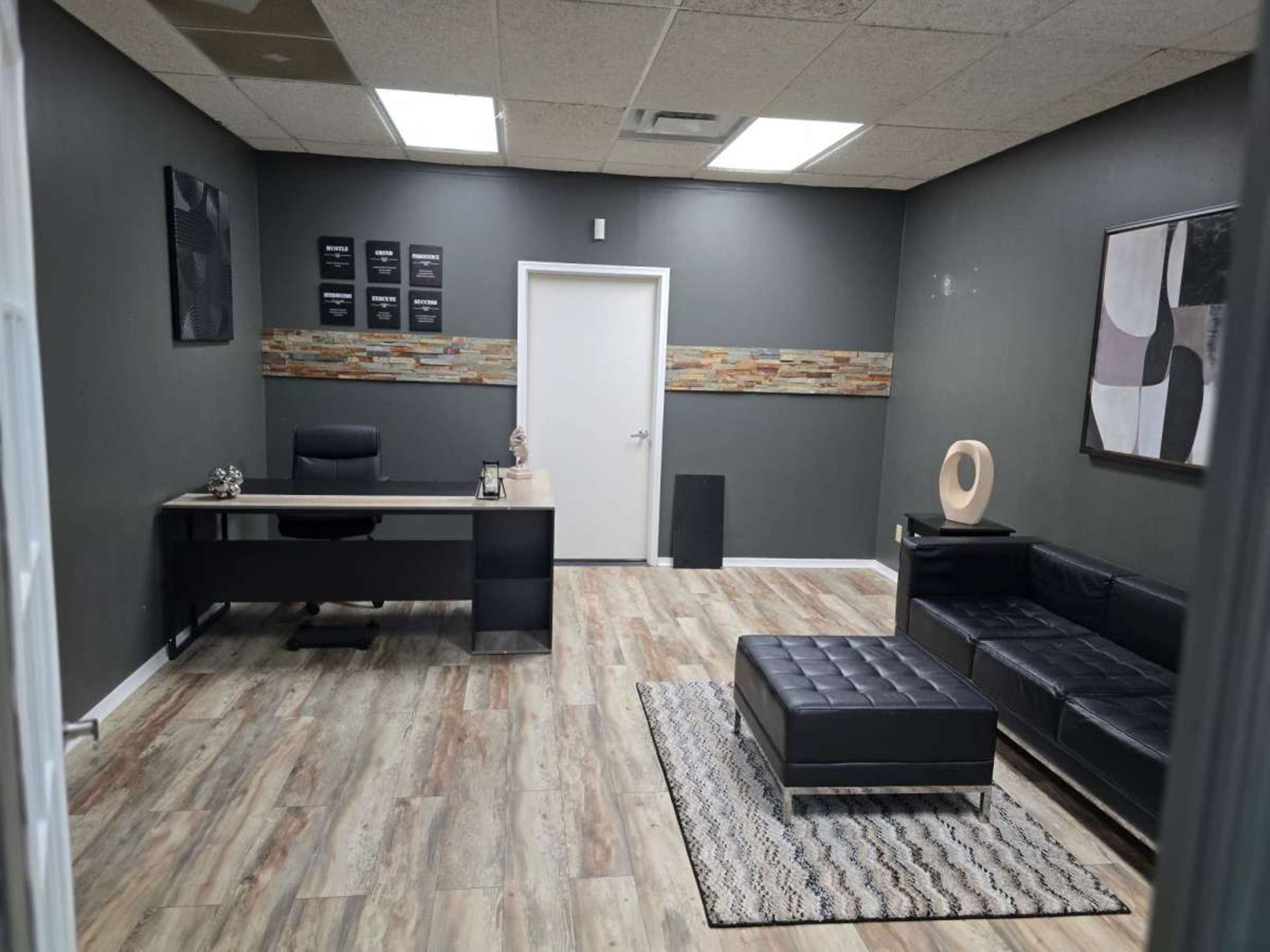 The image shows a modern, minimalist office space featuring a black desk with a chair, a black couch, and a decorative rug on a wooden floor.