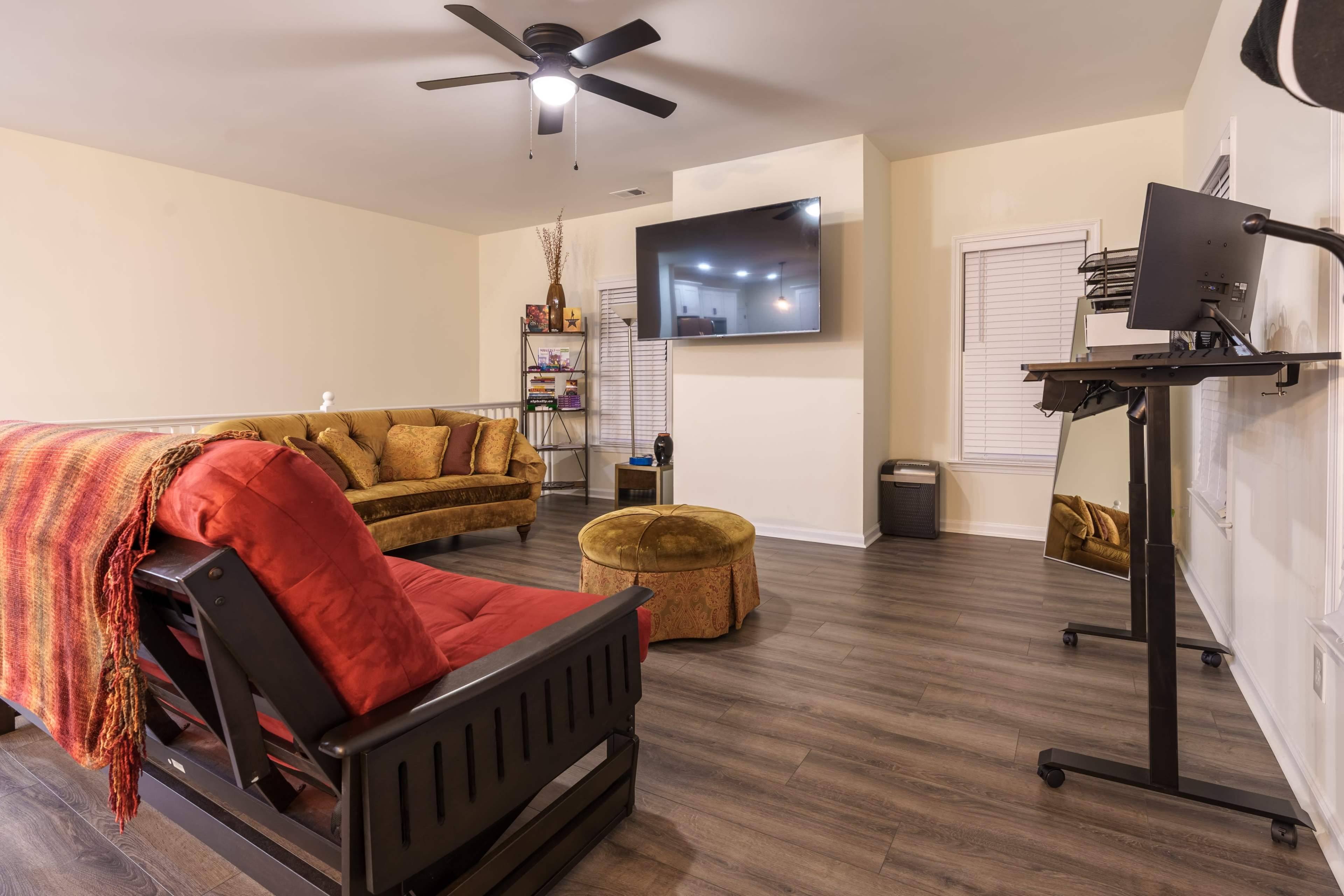 A living room features a brown sofa, a red chair, an ottoman, a television mounted on the wall, and a standing desk in a corner.