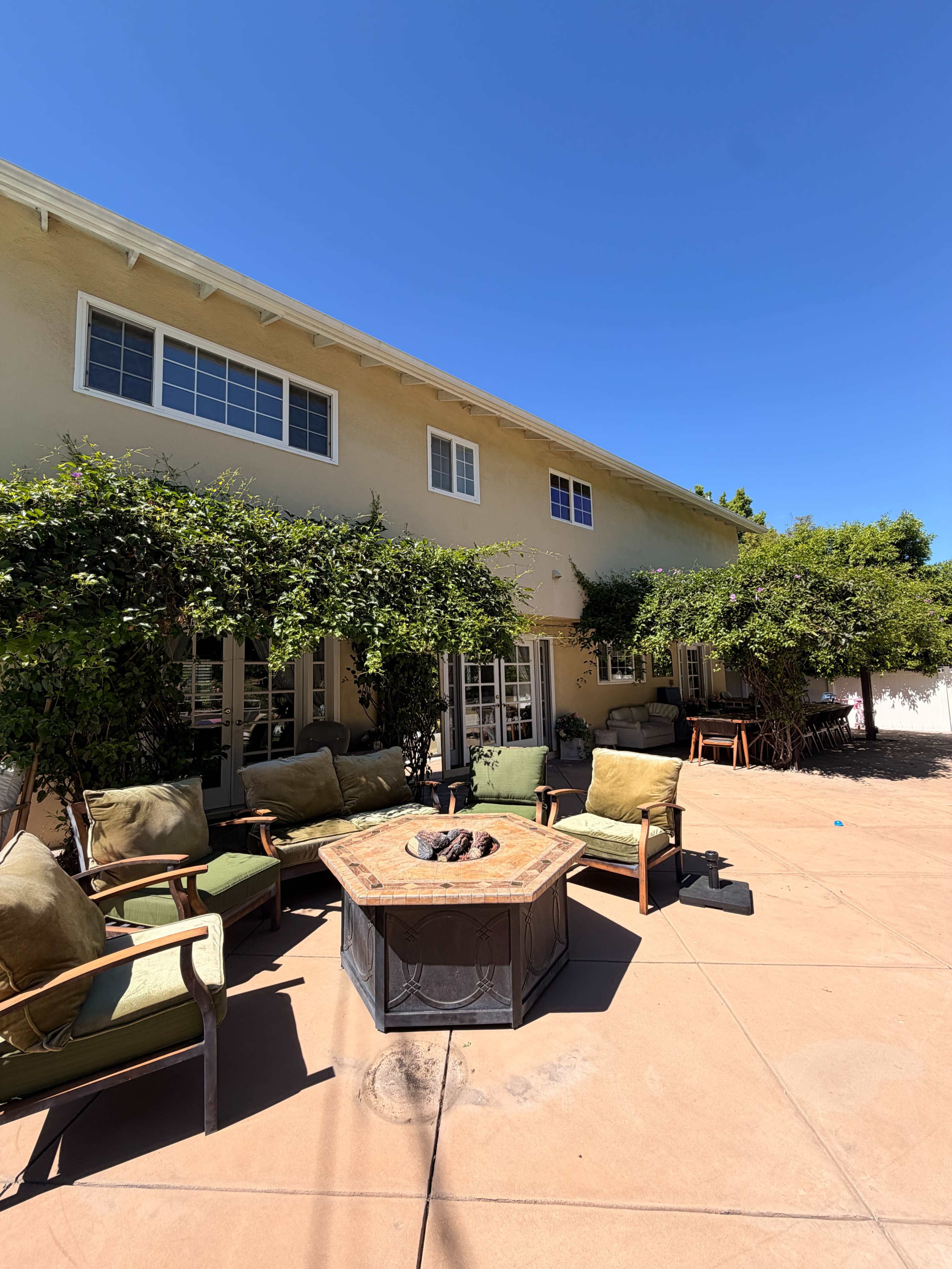 A patio area features a fire pit surrounded by chairs, with a two-story house and lush greenery in the background under a clear blue sky.