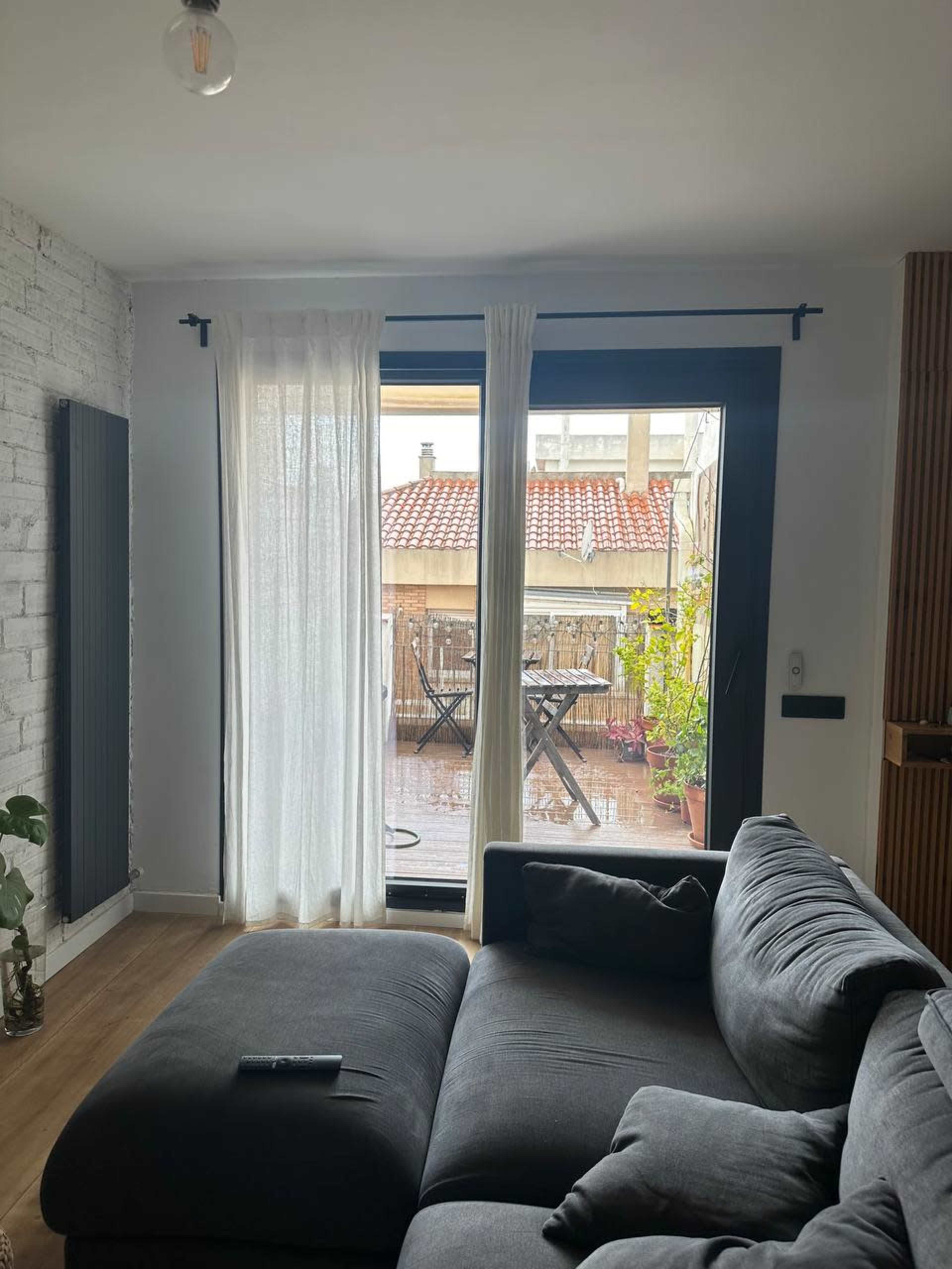 A gray sectional sofa faces a large window with sheer curtains, revealing a rainy balcony with a dining table and potted plants.