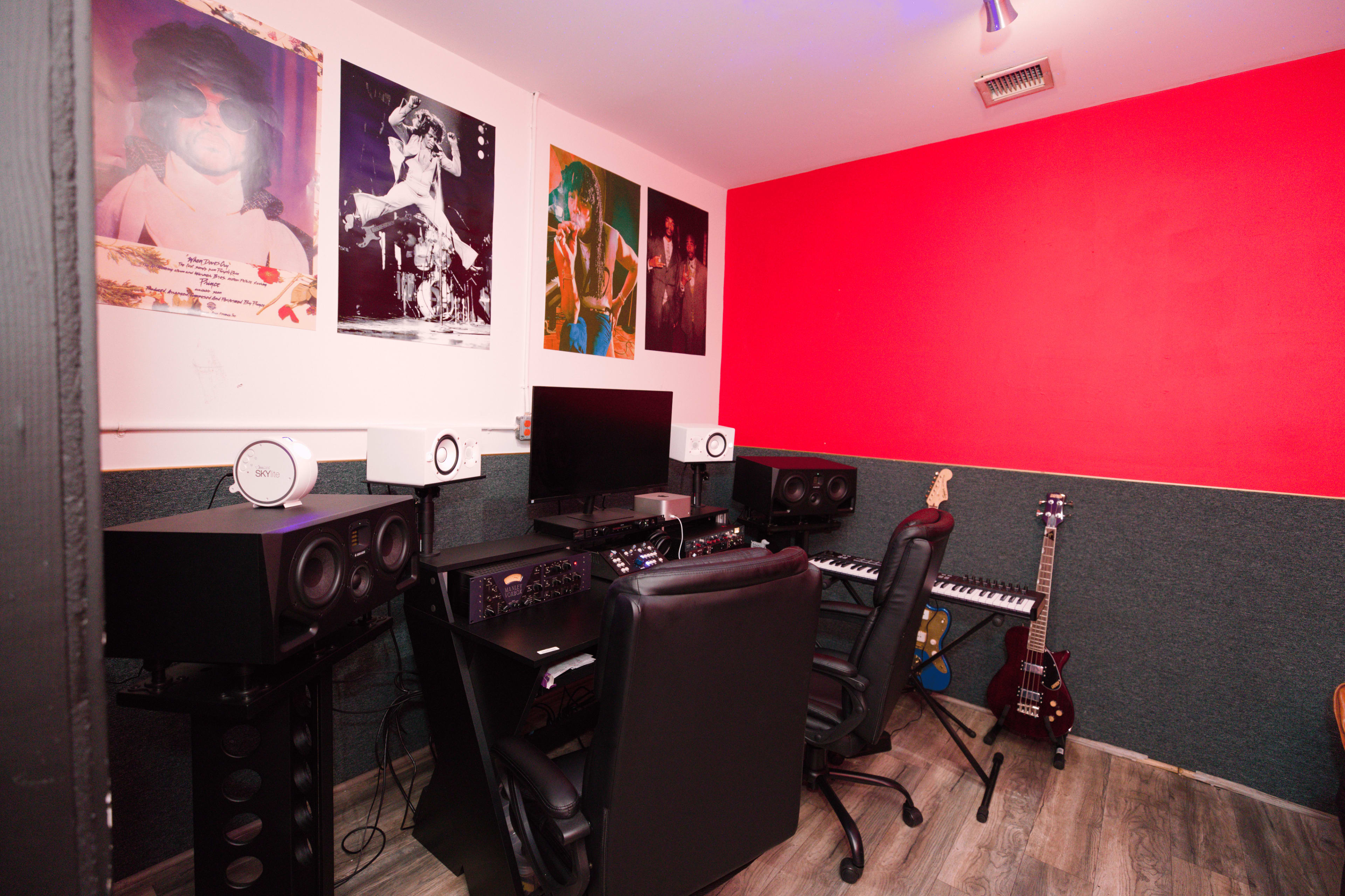 The image shows a music production studio with two computer desks, studio monitors, a keyboard, and various posters on the walls.