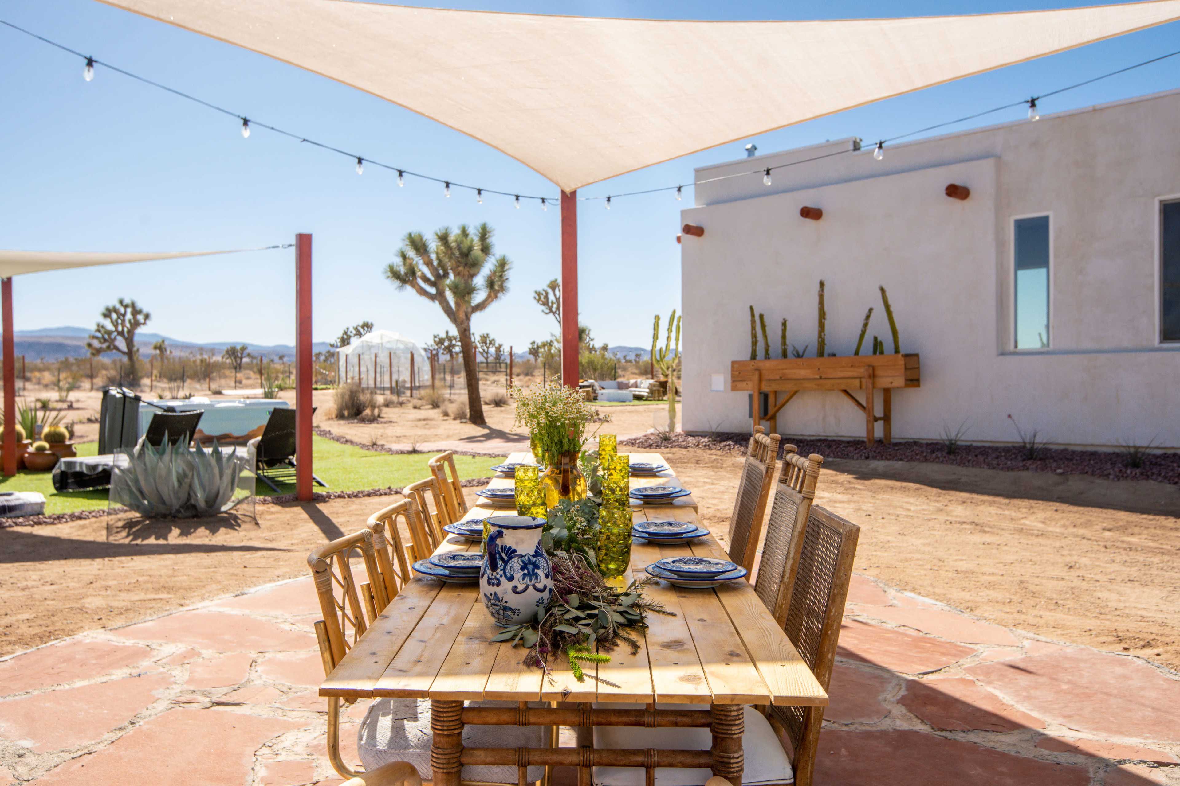 A wooden dining table is set outdoors under a shaded area with a desert landscape and cacti in the background.