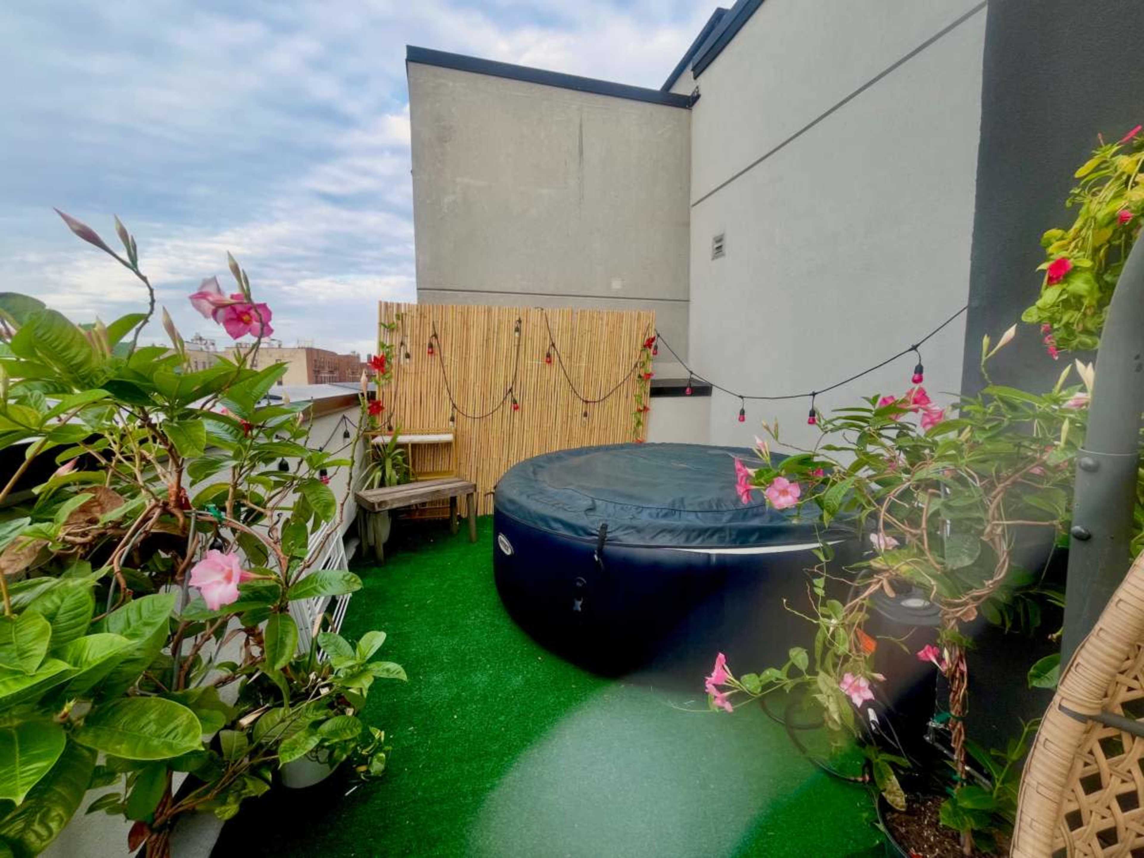 The image shows a rooftop terrace with a circular hot tub surrounded by potted plants and decorative string lights.