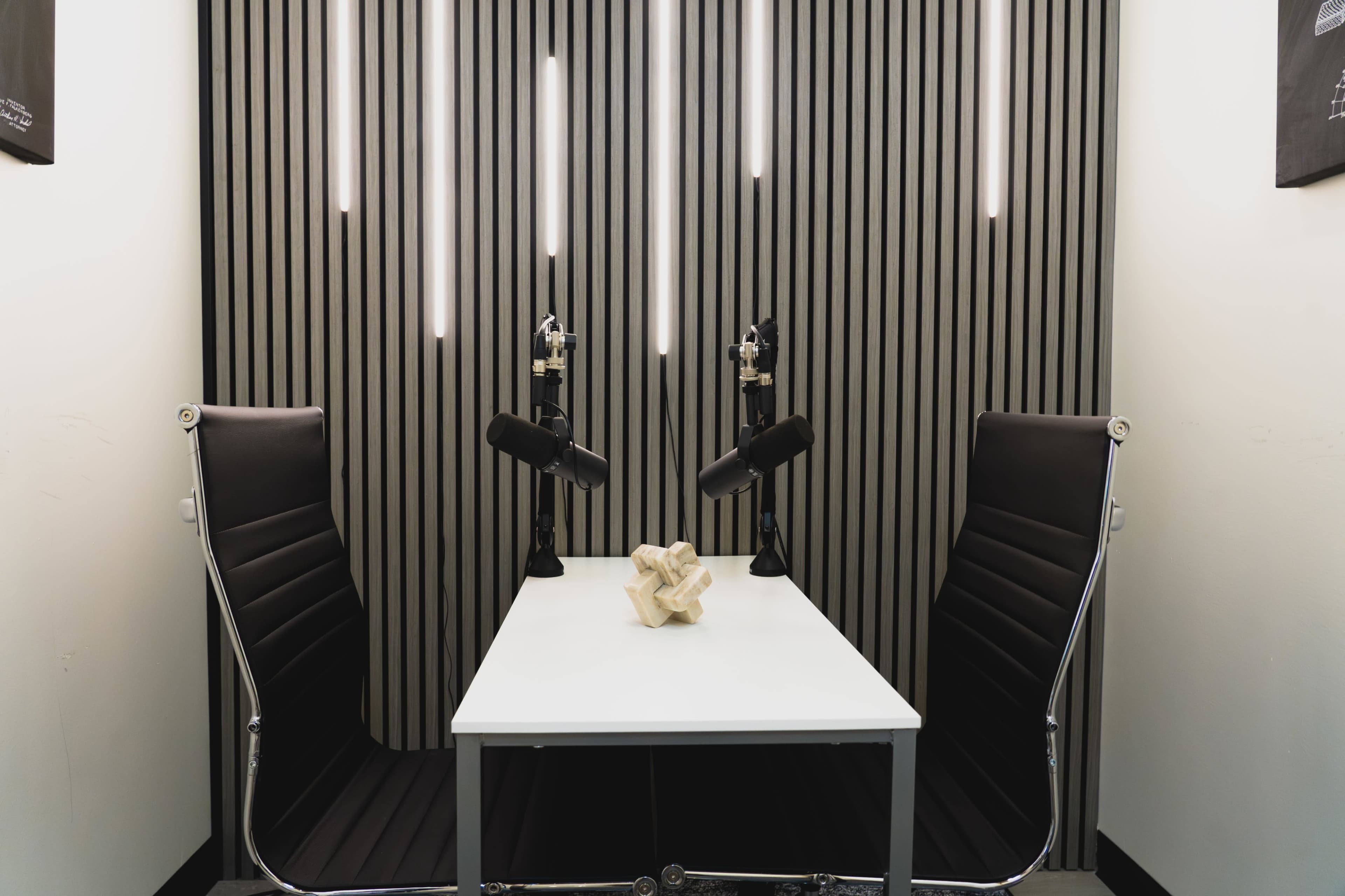 The image shows a podcast recording studio featuring a white table, two black leather chairs, and two microphones mounted on stands against a striped wall illuminated by overhead lights.