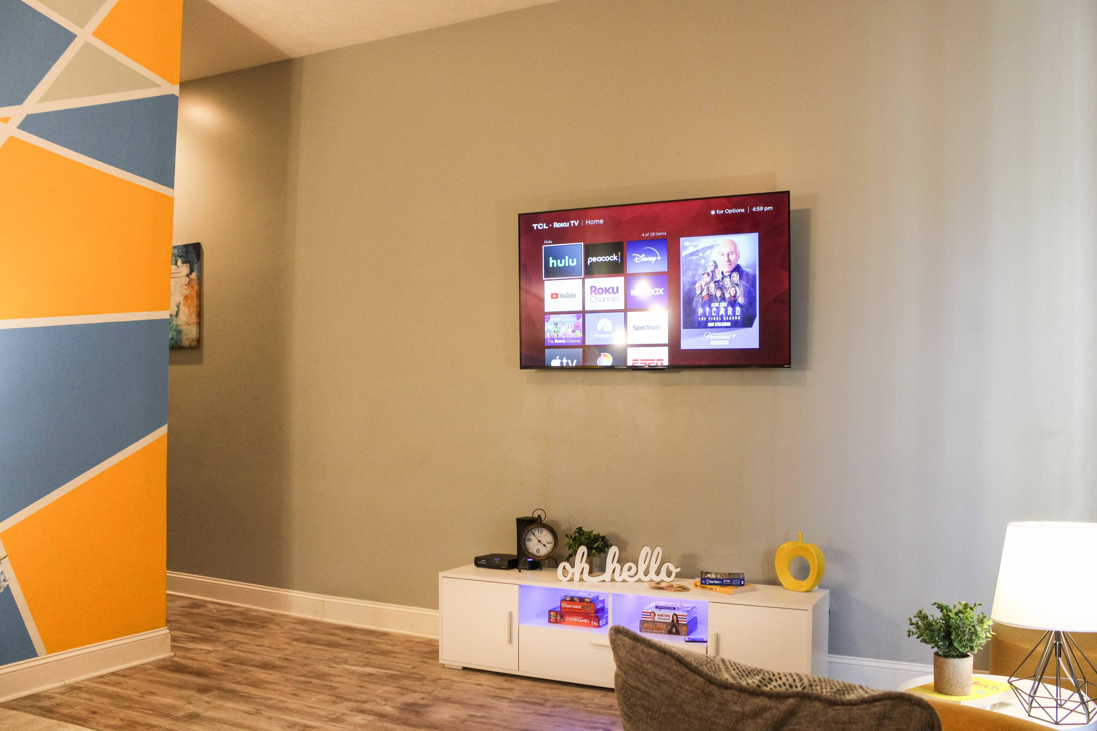 A living room features a television mounted on the wall displaying streaming service options, with a decorative console beneath it.