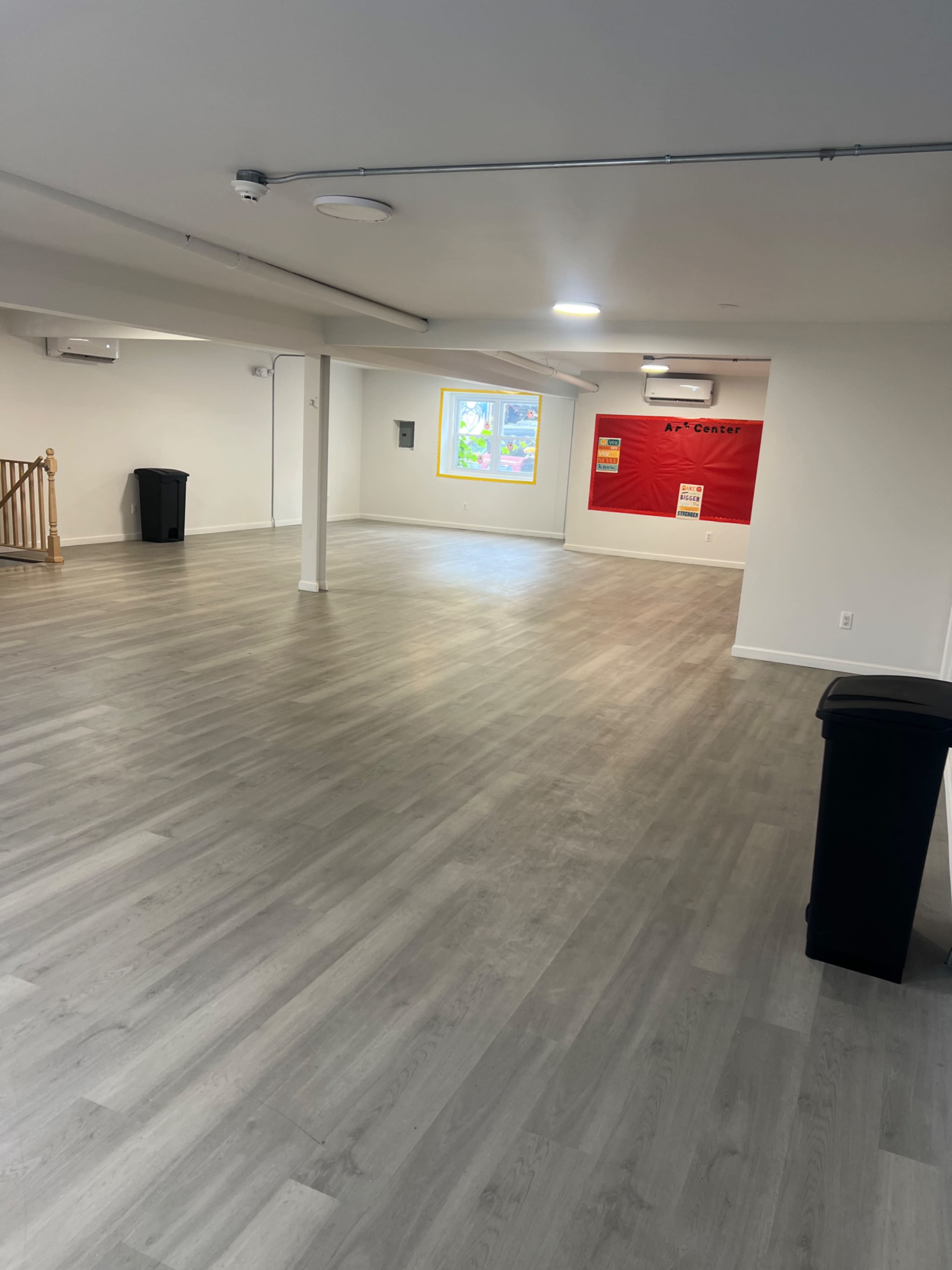 The image shows a spacious, empty room with light gray flooring, a trash bin, and a wall featuring a red accent with posters.