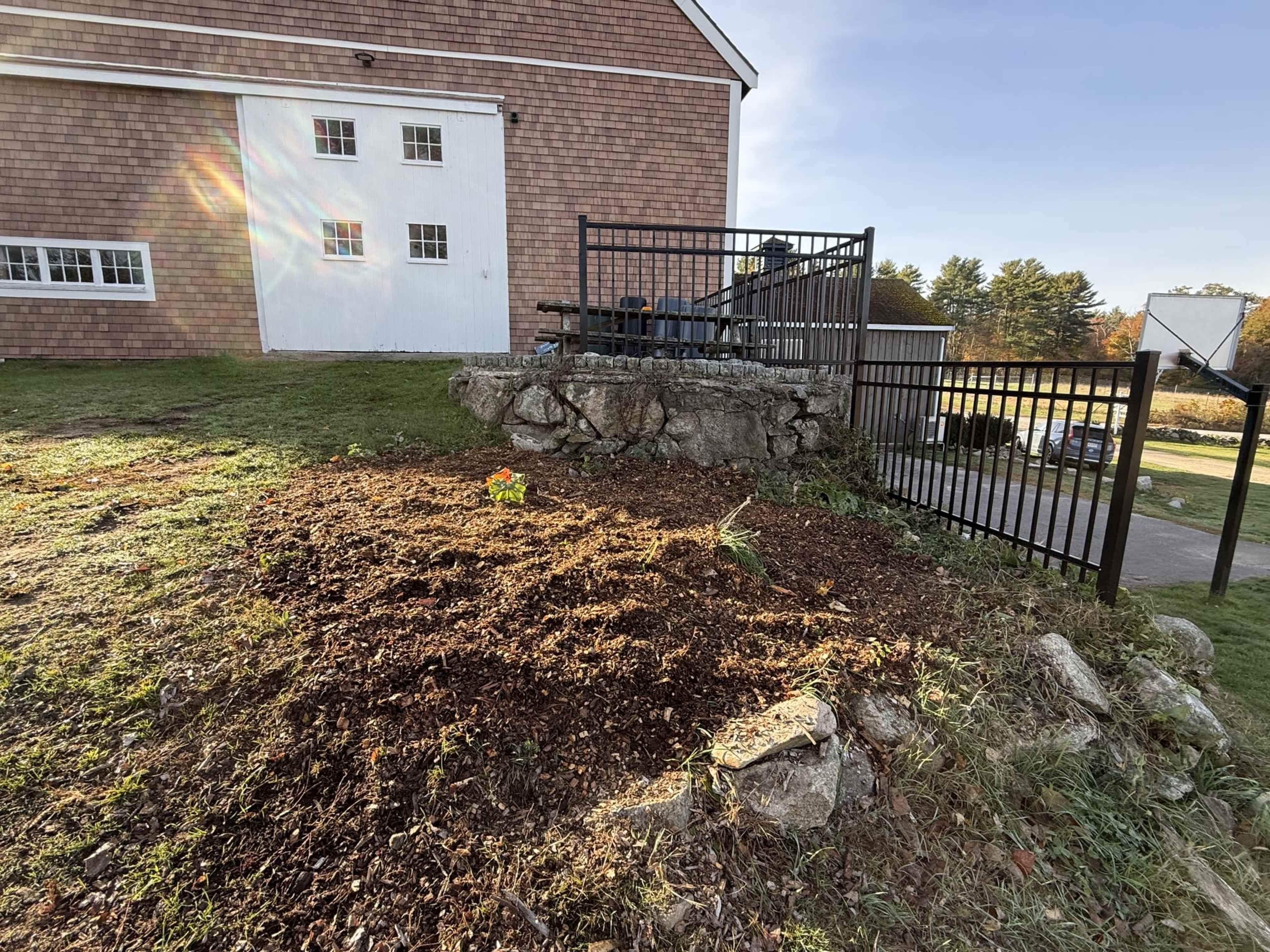 The image shows a grassy area with a mulch-covered patch and a rock formation next to a fenced in platform and a building.