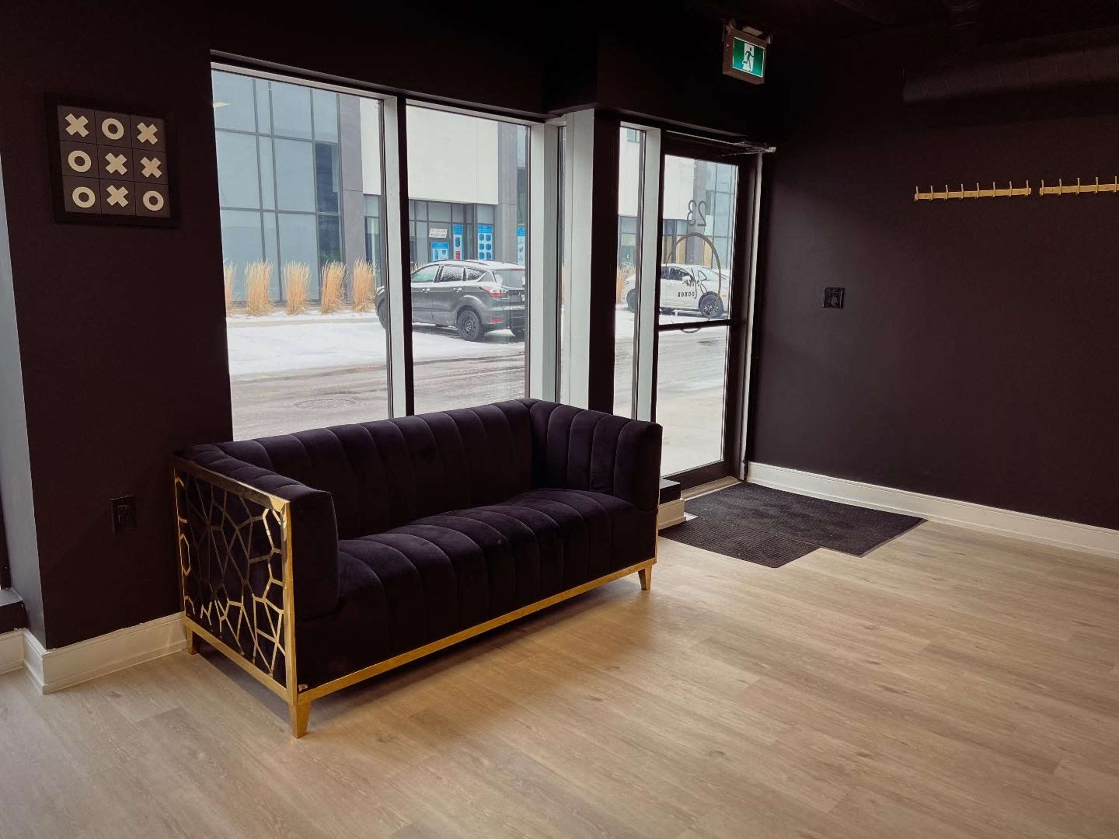The image shows a modern, dark-toned interior featuring a black sofa with gold accents positioned near a glass entrance, with snow visible outside.