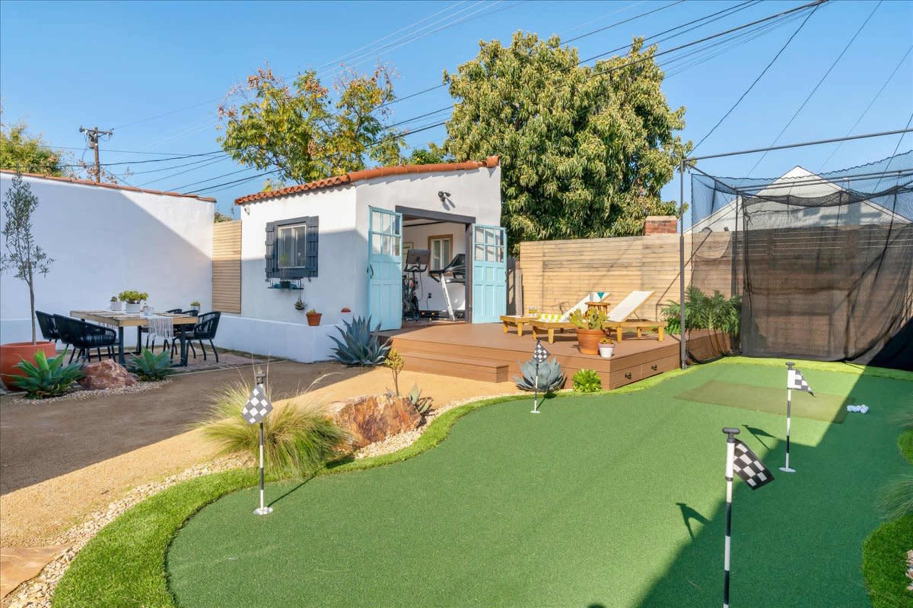 The image shows a landscaped backyard featuring a green putting green, an outdoor seating area with a table, a small house with blue doors, and a netted sports area.
