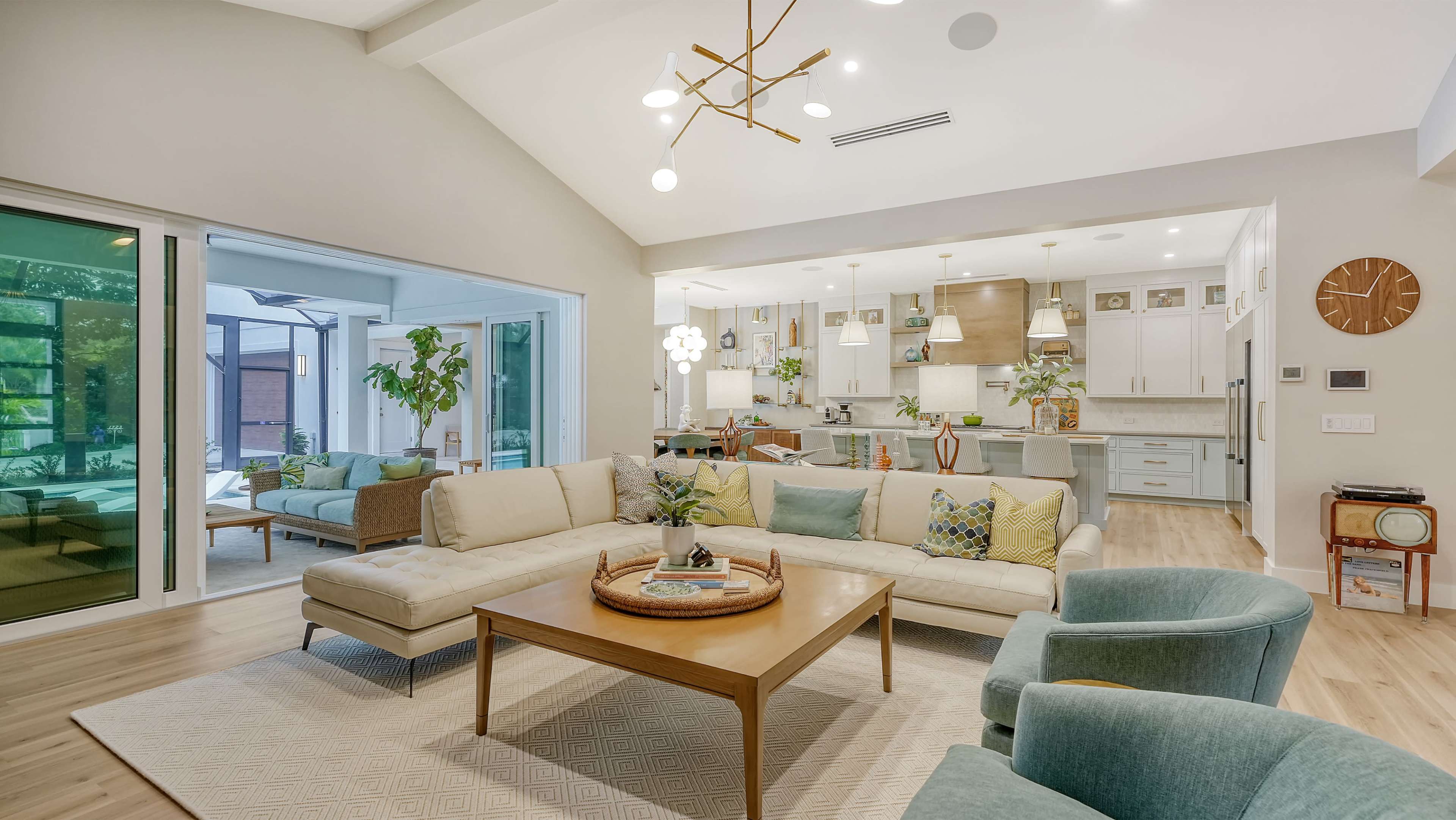 The image depicts a modern living room with a sectional sofa, a wooden coffee table, and a view into a kitchen featuring light cabinetry and multiple plants throughout the space.