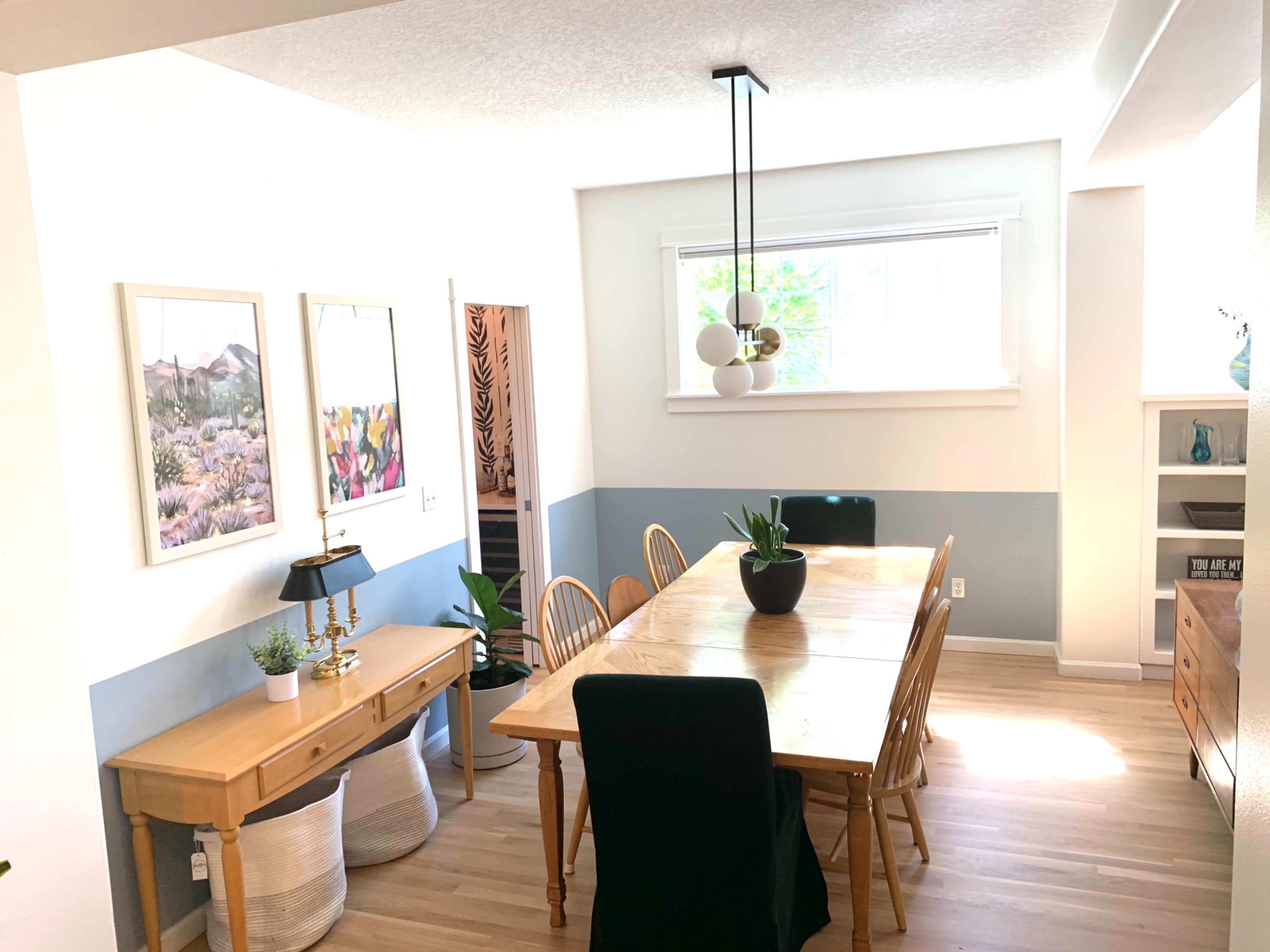 A dining area with a wooden table surrounded by chairs, a console table with a lamp, and framed artwork on the walls.